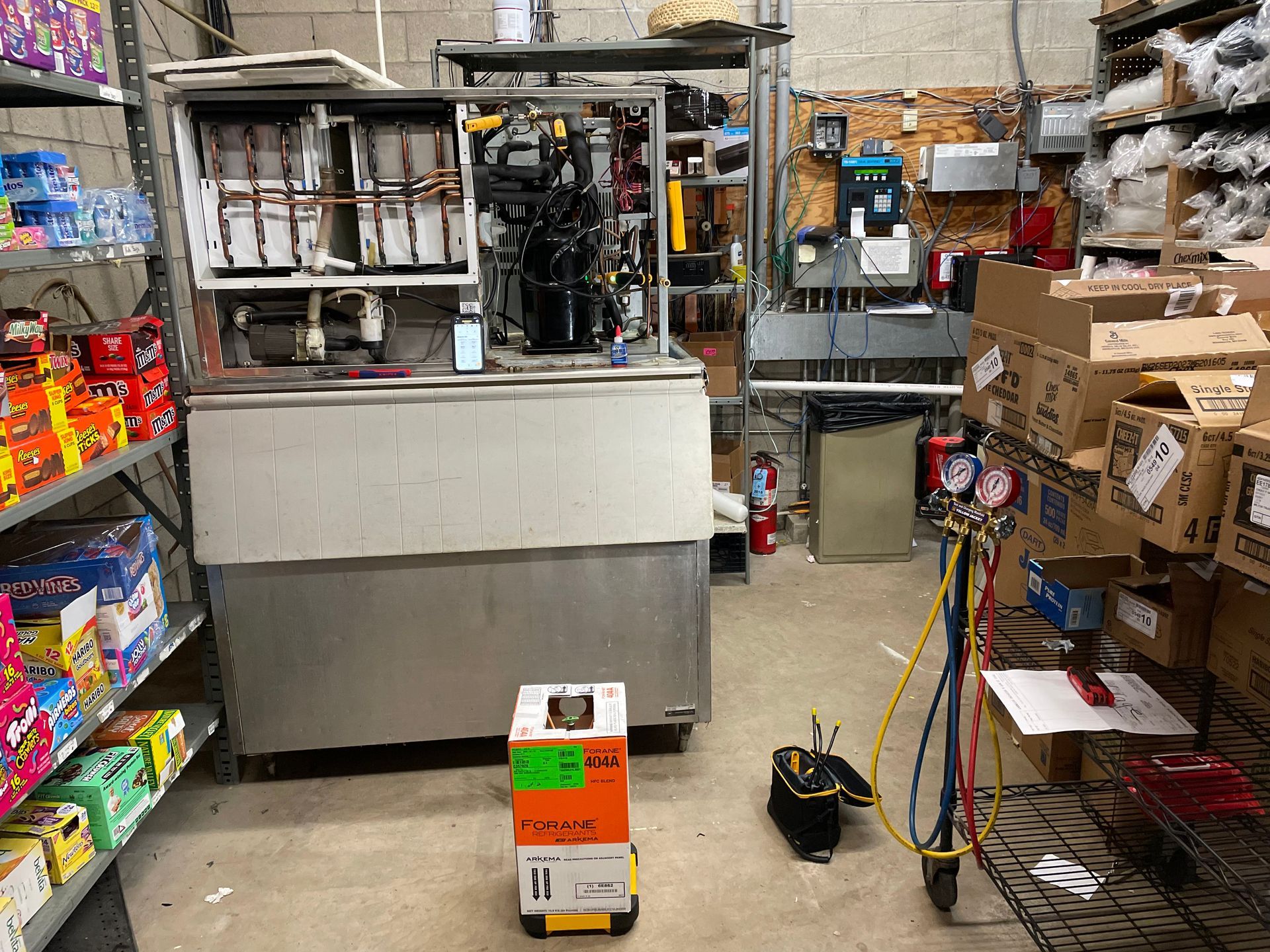 Inside a cluttered store, a refrigeration unit is being worked on. Gauges, tools, and boxes are present.