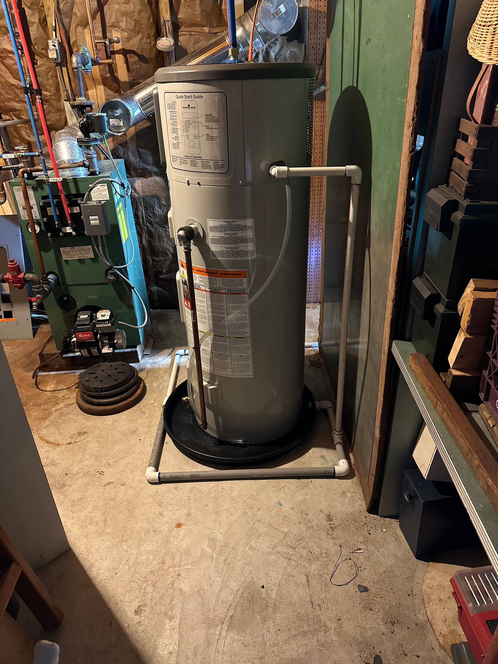 Large gray water heater in a basement utility area, surrounded by pipes and tools.