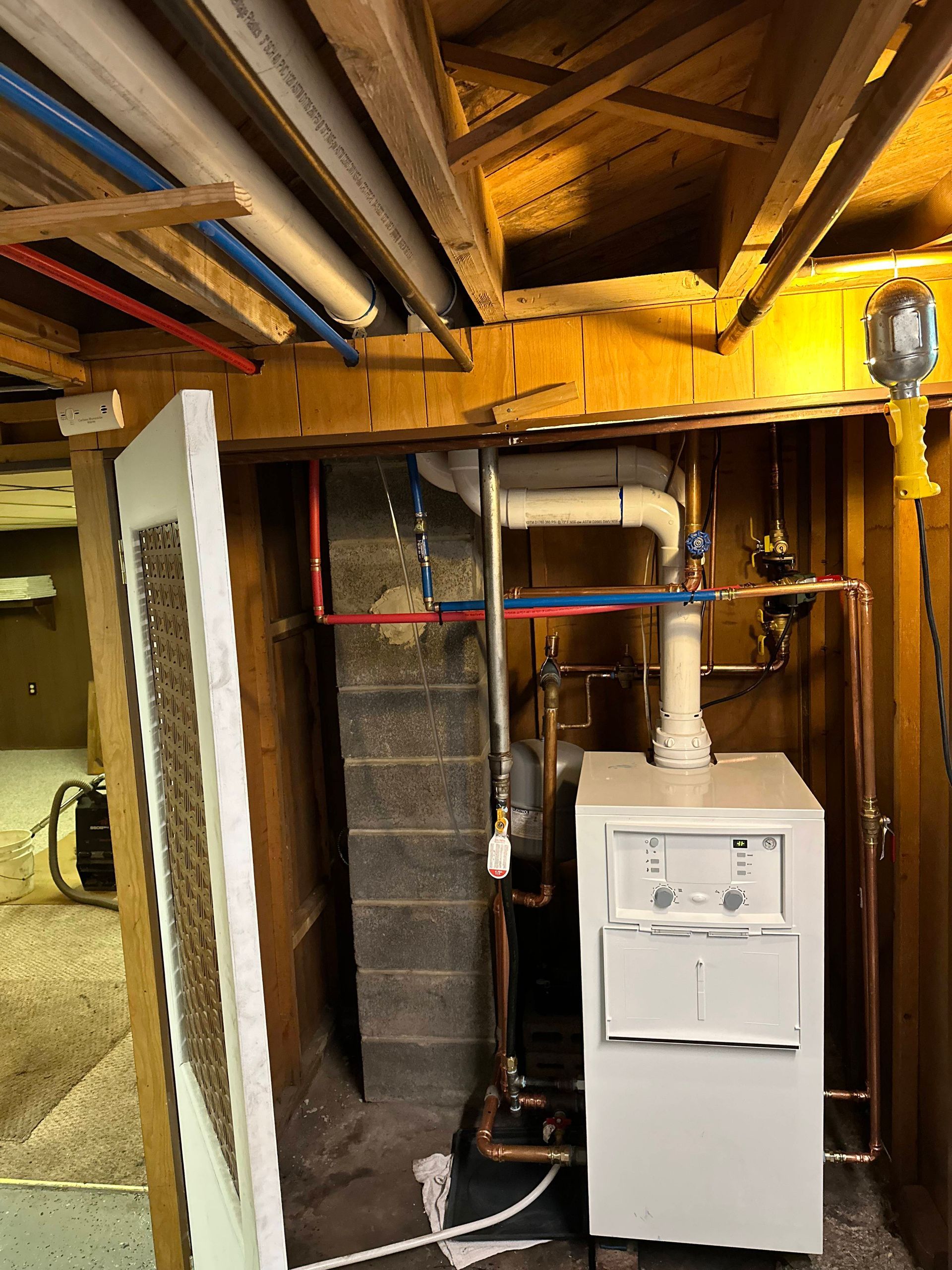 Basement utility area with a white furnace, exposed pipes, and wooden ceiling beams