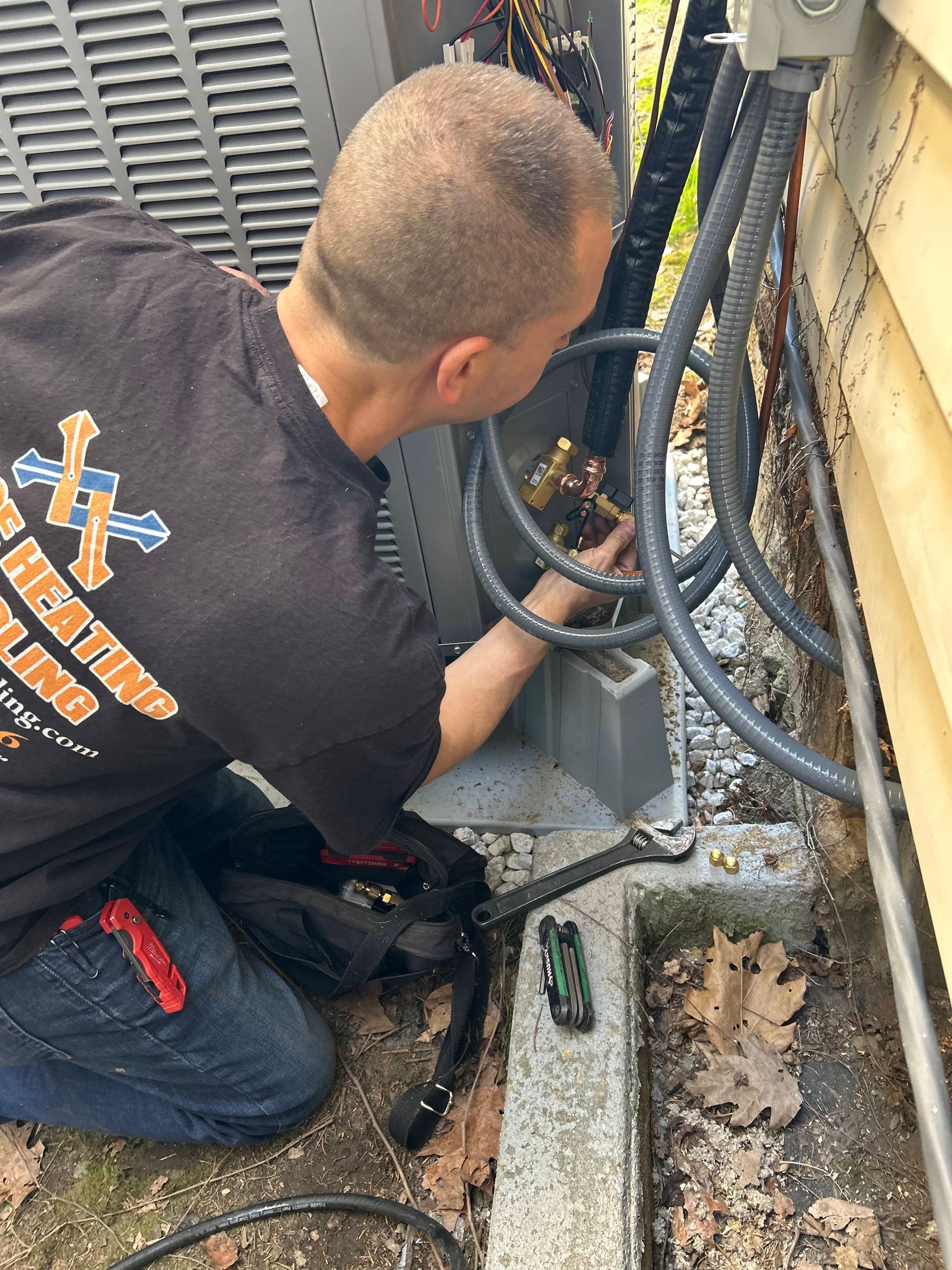 Technician kneels by outdoor cables, inspecting wiring near an HVAC unit.