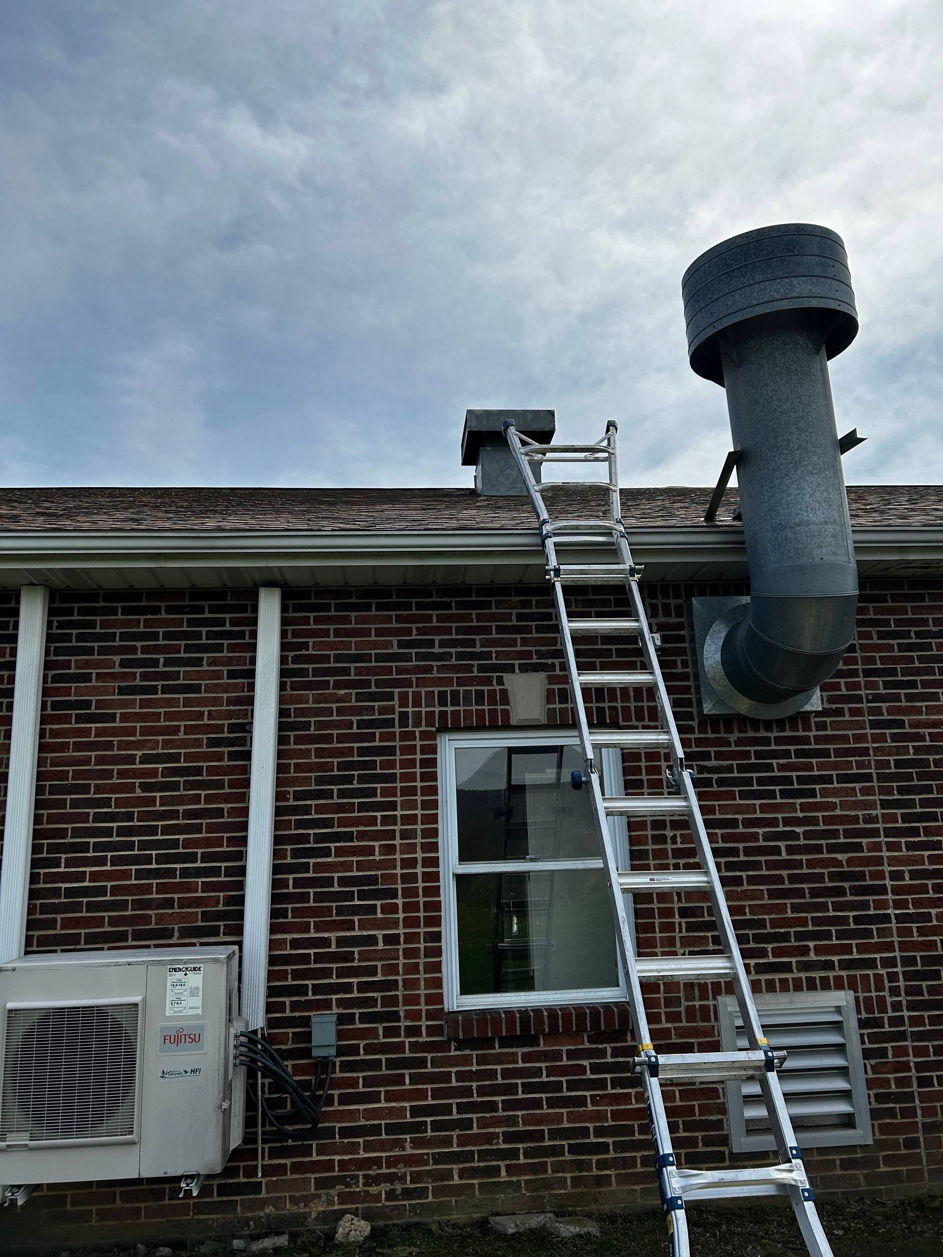 Ladder leaning against a brick house roof, with a chimney vent and an air conditioner below.