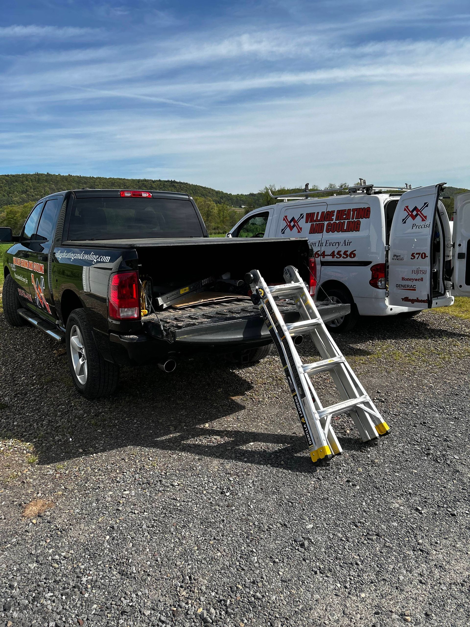 Black SUV with open hatch and rear ramp on gravel beside a white service van under a blue sky.