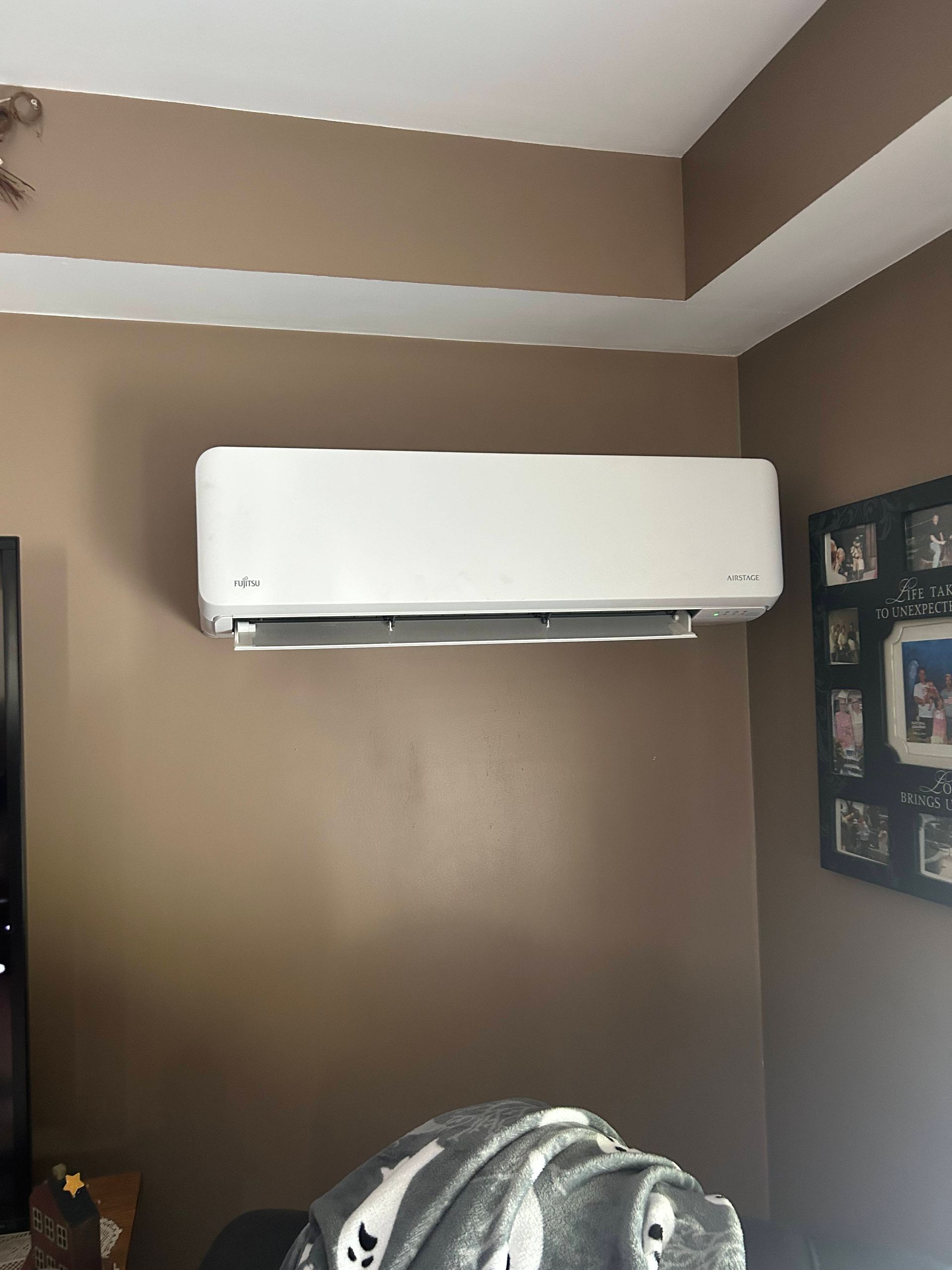 White wall-mounted air conditioner on a brown wall in a room corner, with framed photos nearby.
