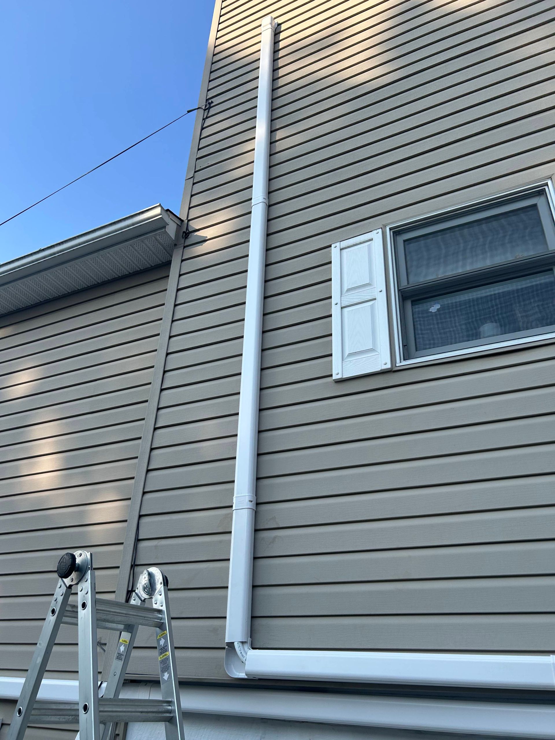 Ladder against tan house siding with white trim and downspout beneath a blue sky