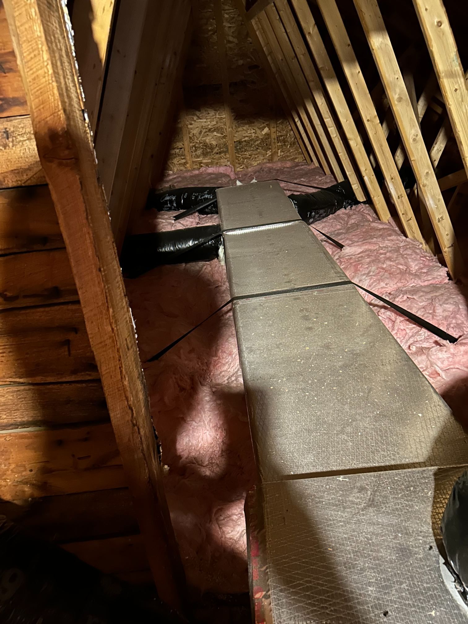 Narrow attic corridor with exposed wooden rafters and a concrete walkway on pink insulation.