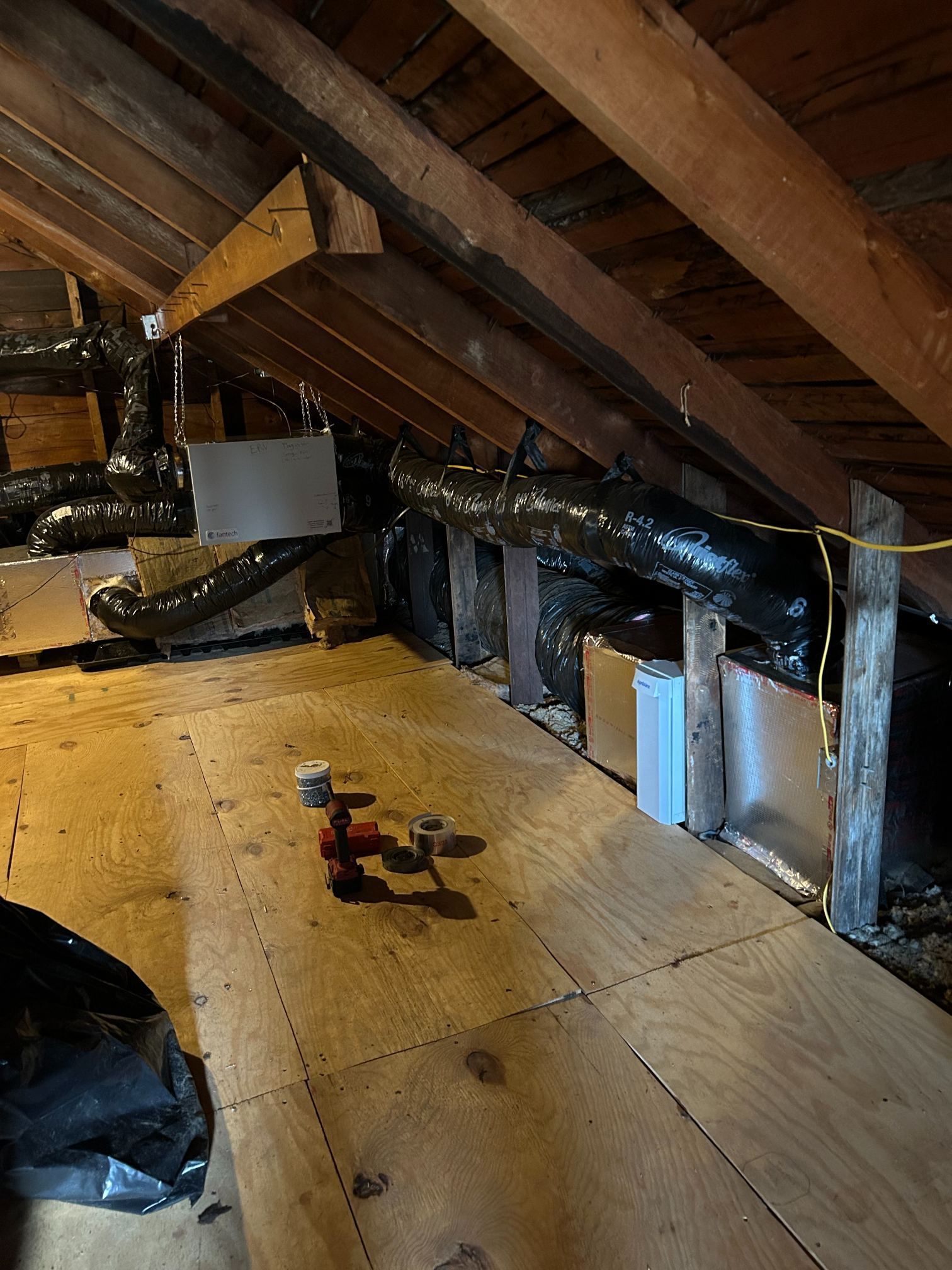 Attic with exposed rafters, plywood floor, and insulation, with a small red drill on the floor.