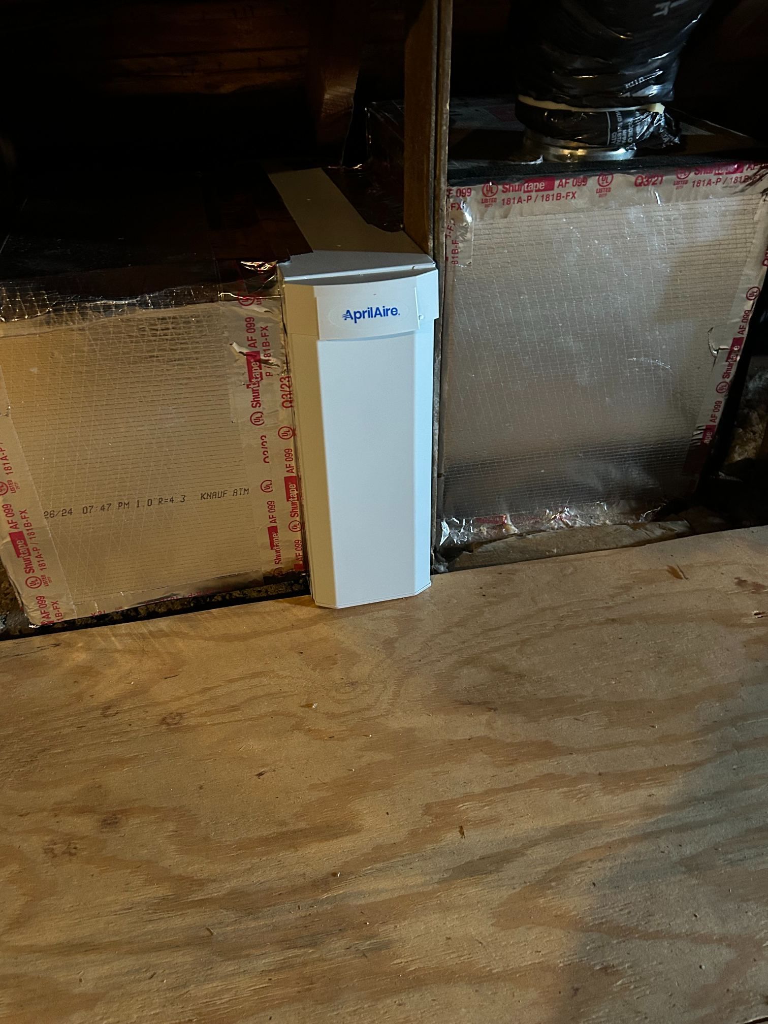 White rectangular device standing on a wooden floor beside insulated ductwork in an attic space