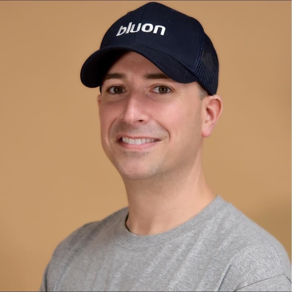 Man wearing a navy blue baseball cap and gray shirt smiles in front of a tan background.