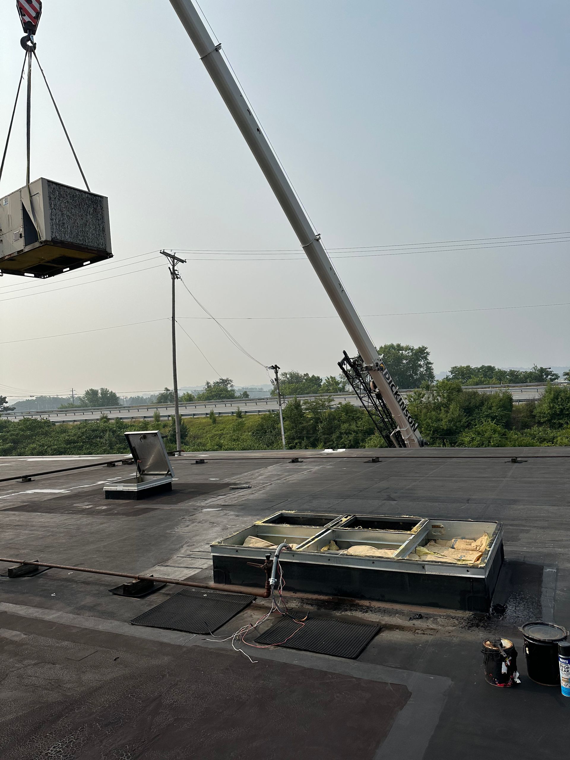 Crane lifting a large HVAC unit onto a flat roof with vents, a pole, and a cityscape in the background.