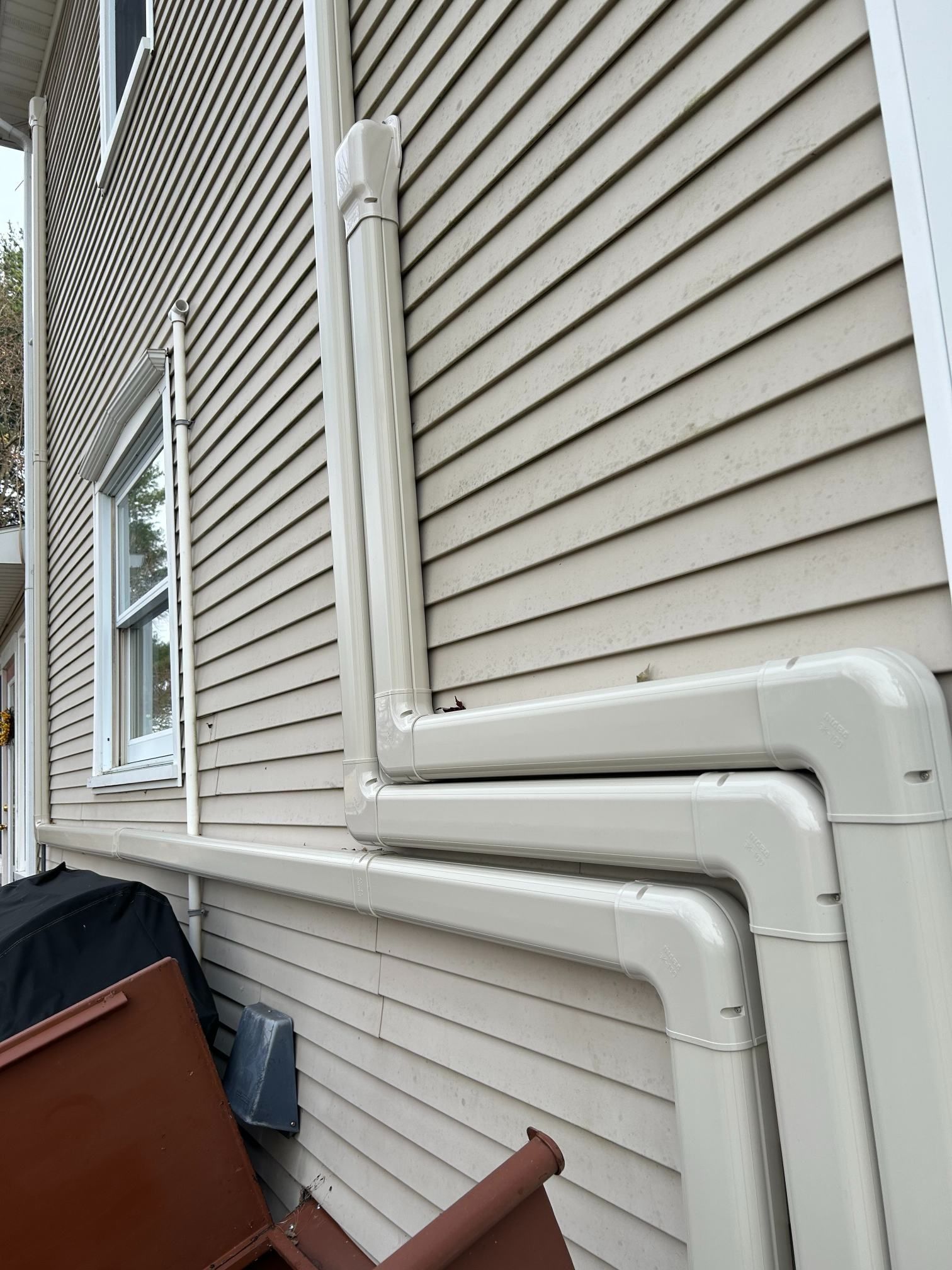 Exterior house wall with beige siding and white drain pipes running along the side near a window.