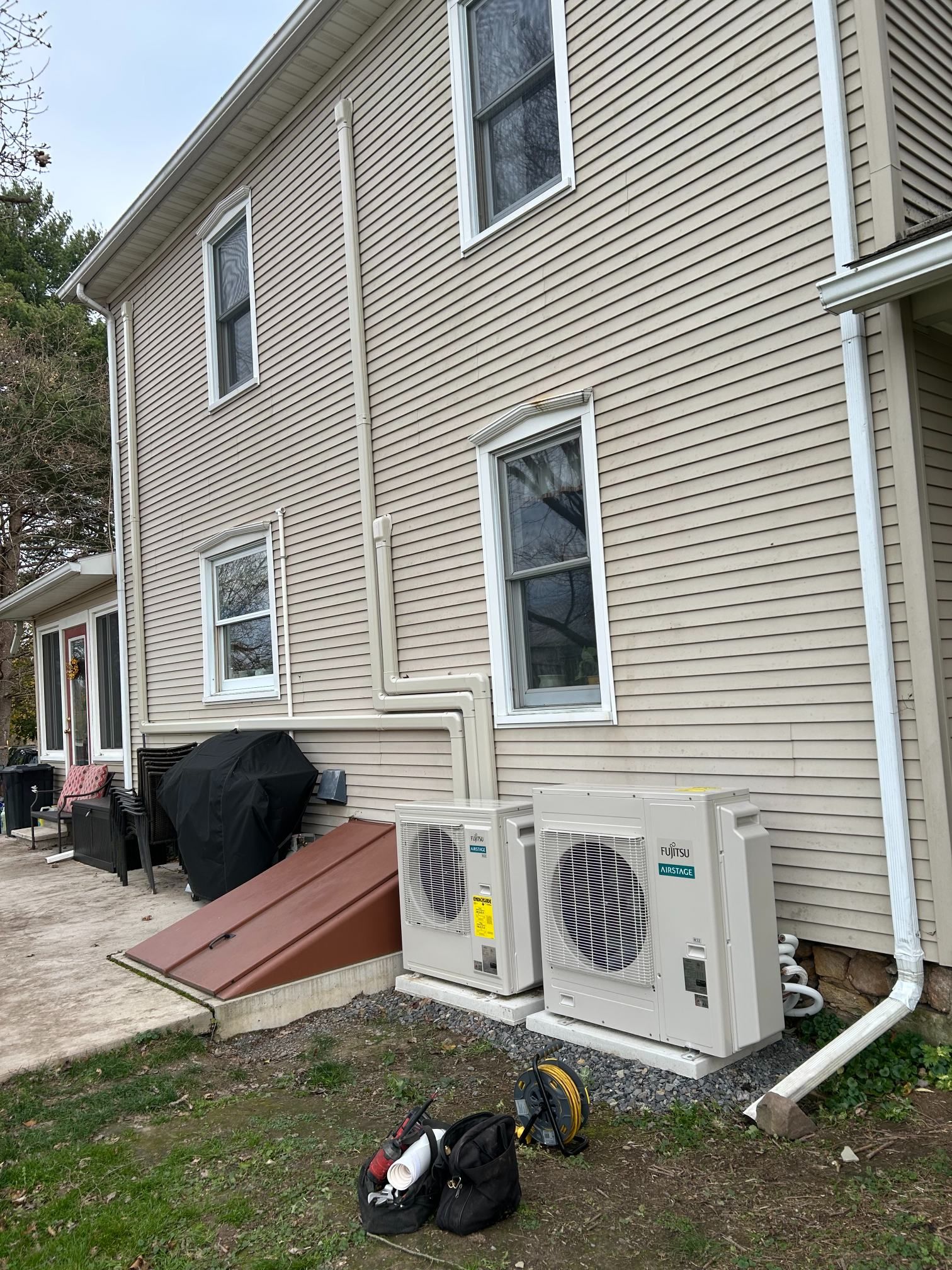 Side of beige house with air conditioners, basement stairs, and tools on a paved walkway.