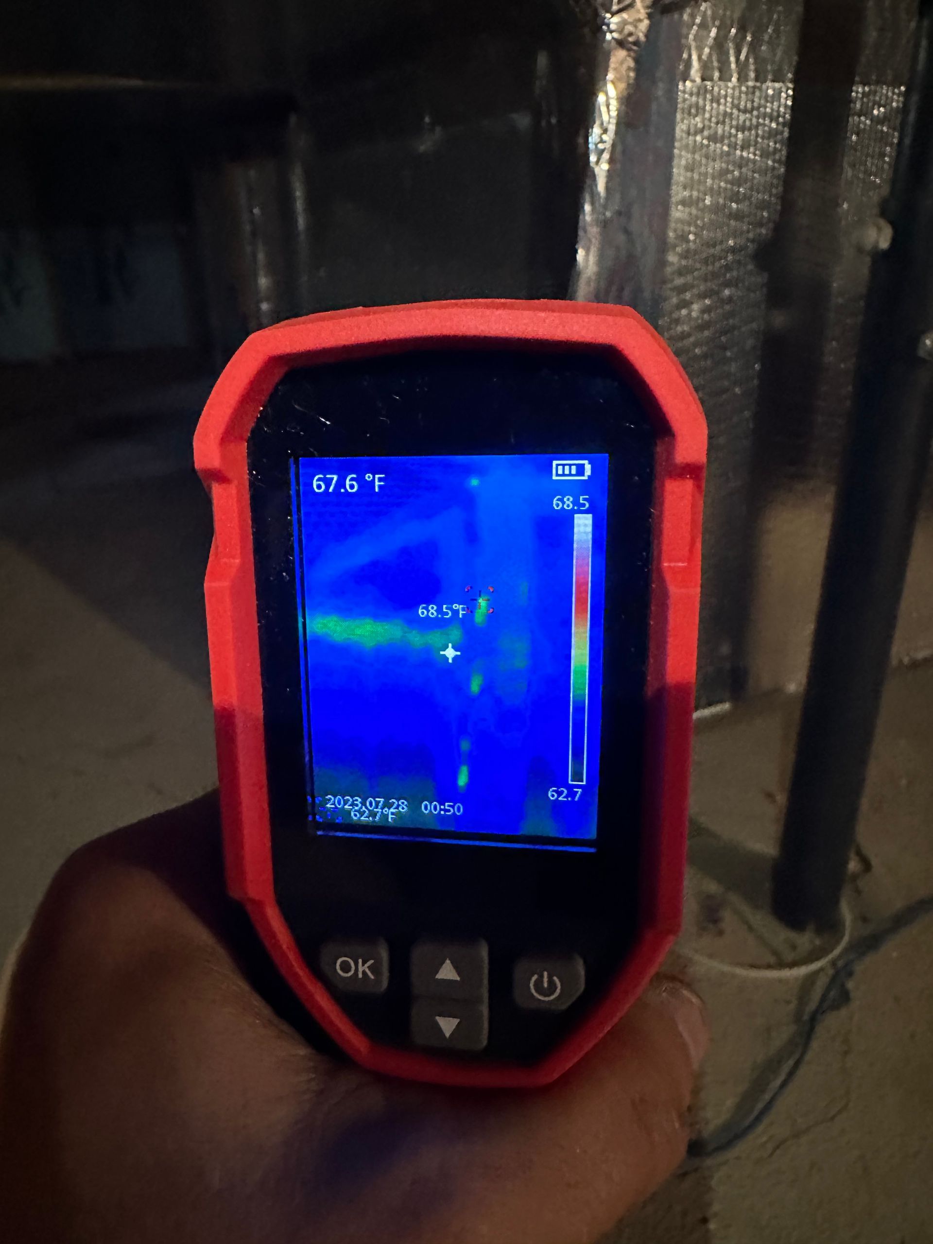 Hand holding thermal camera displaying temperature readings in a dark setting; camera's screen shows blue and green colors.