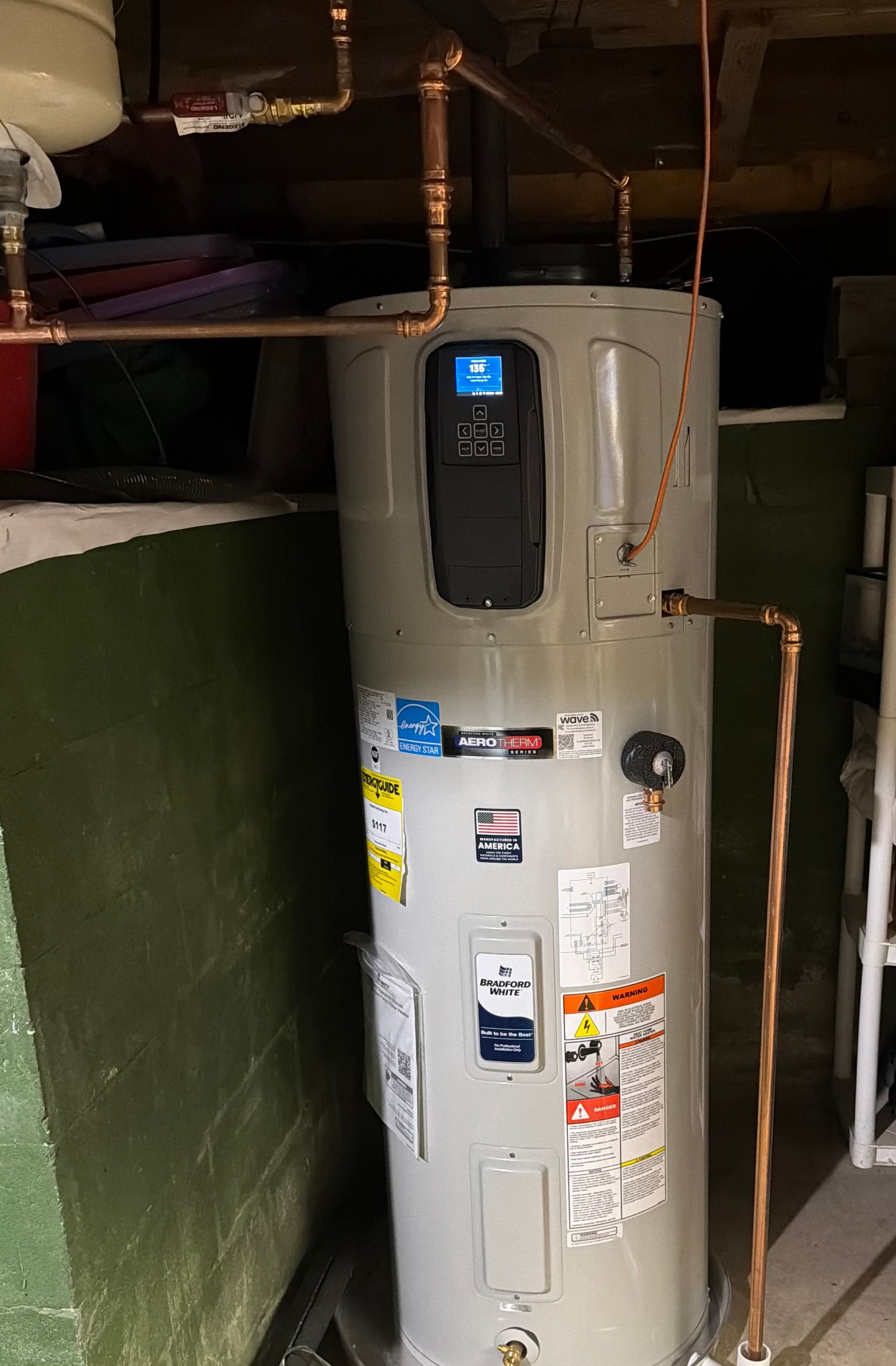Water heater in a basement or utility room with exposed pipes and a small blue display on the front.