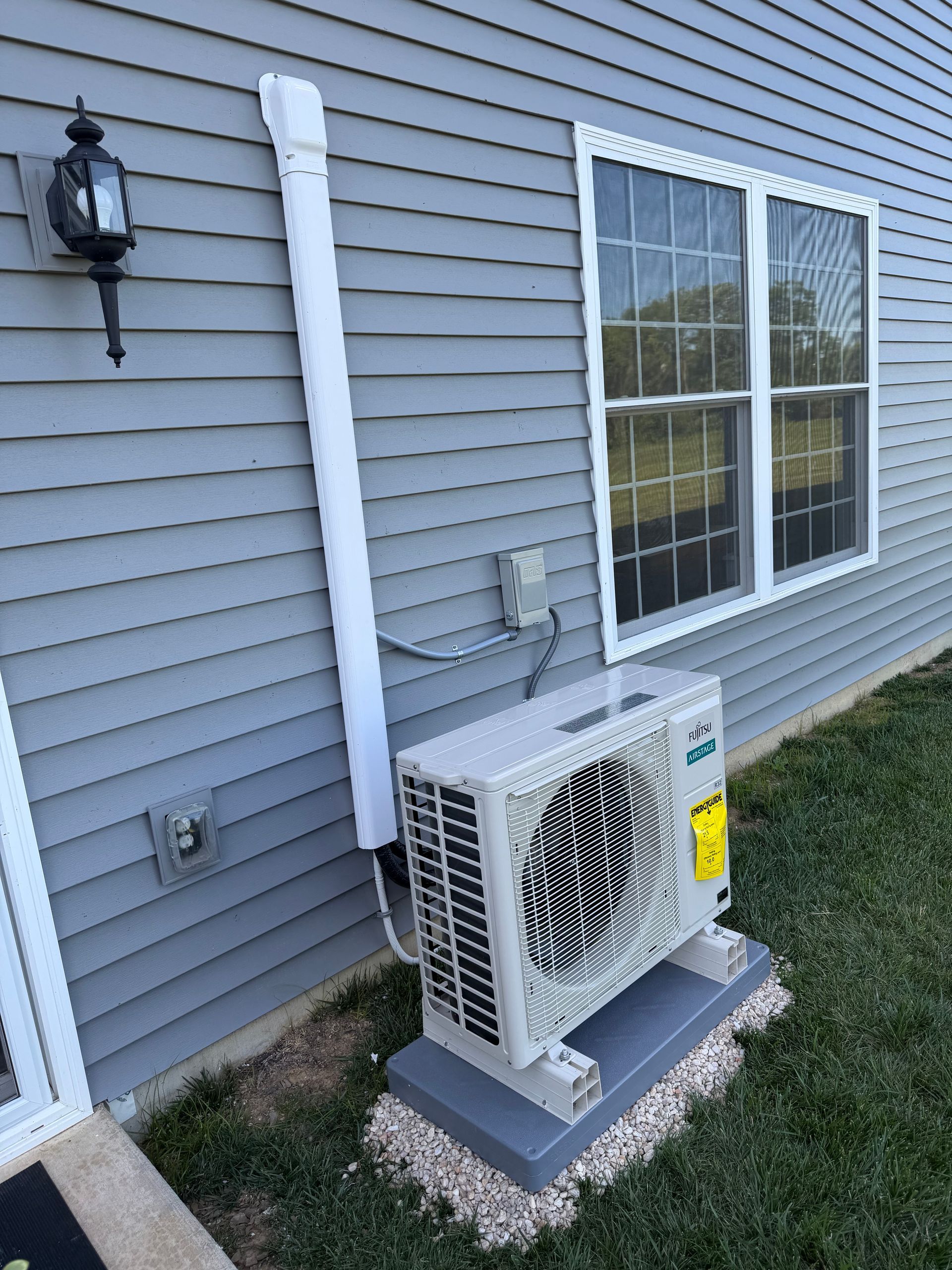 Outdoor air conditioner unit beside a gray house with white trim and window outside wall