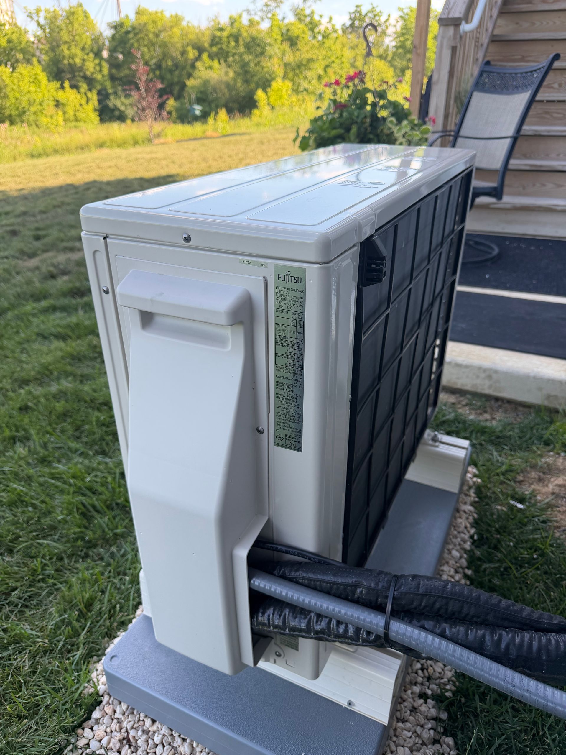 Outdoor HVAC unit beside a house deck, with black hoses attached and a grassy yard in the background