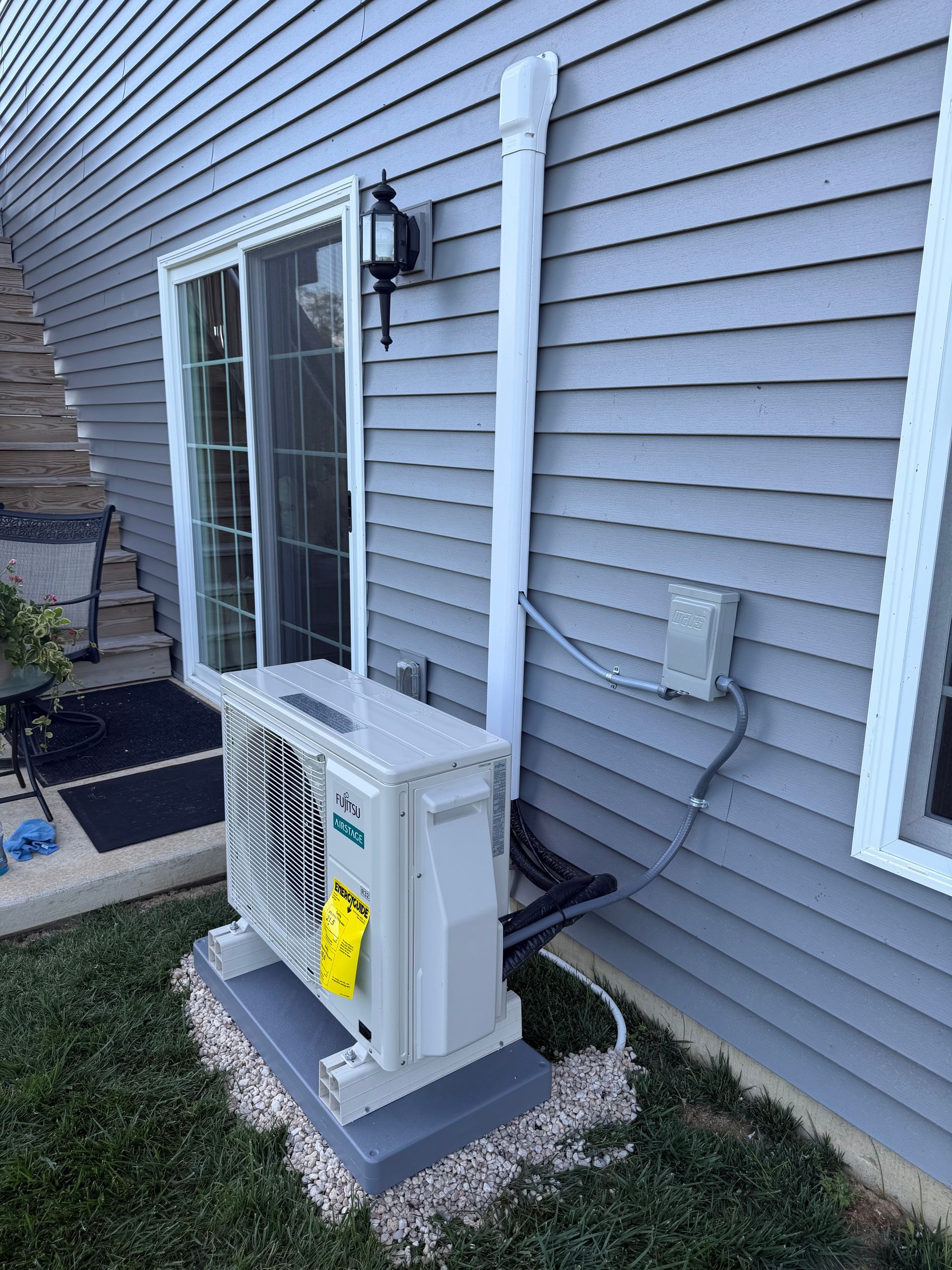 Outdoor HVAC unit beside a gray house wall with a white condensate pipe and window above it