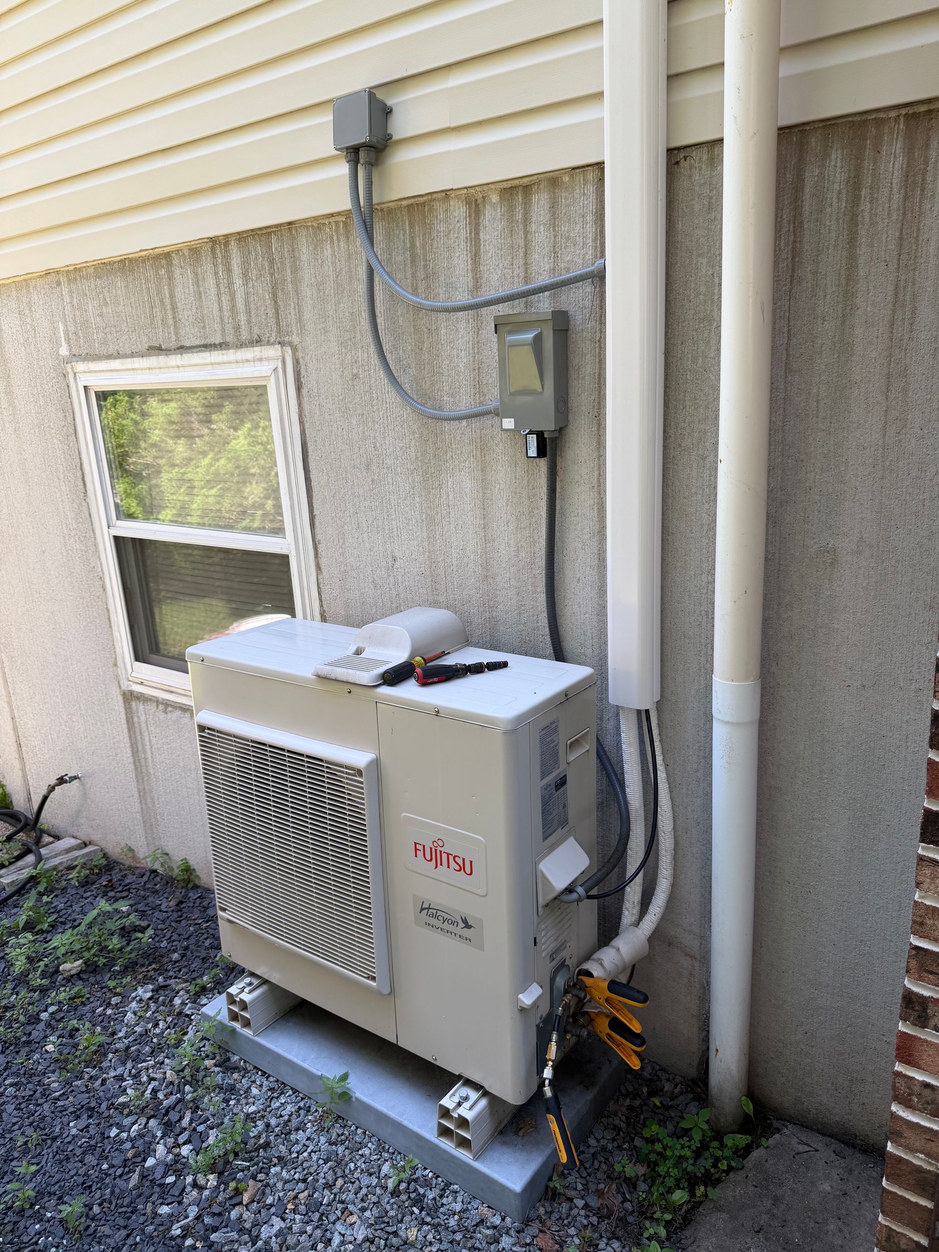 Outdoor air conditioner unit beside a house wall, with pipes and wiring attached.
