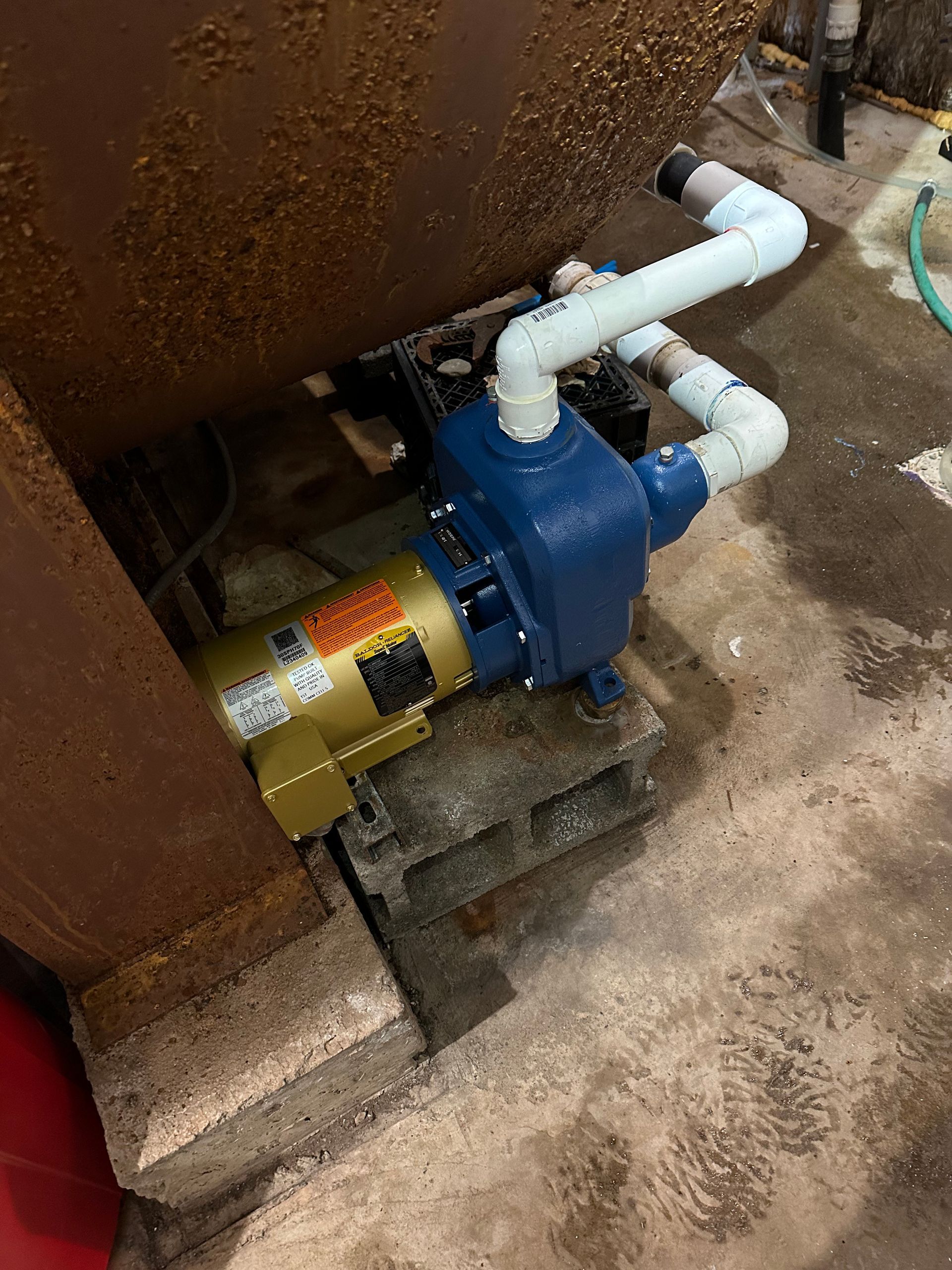 Blue water pump with white PVC pipes, on cinder blocks, near a rusty tank.