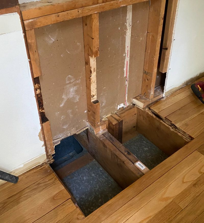 Open section of wall and floor showing wooden frame, possibly for plumbing or electrical work.