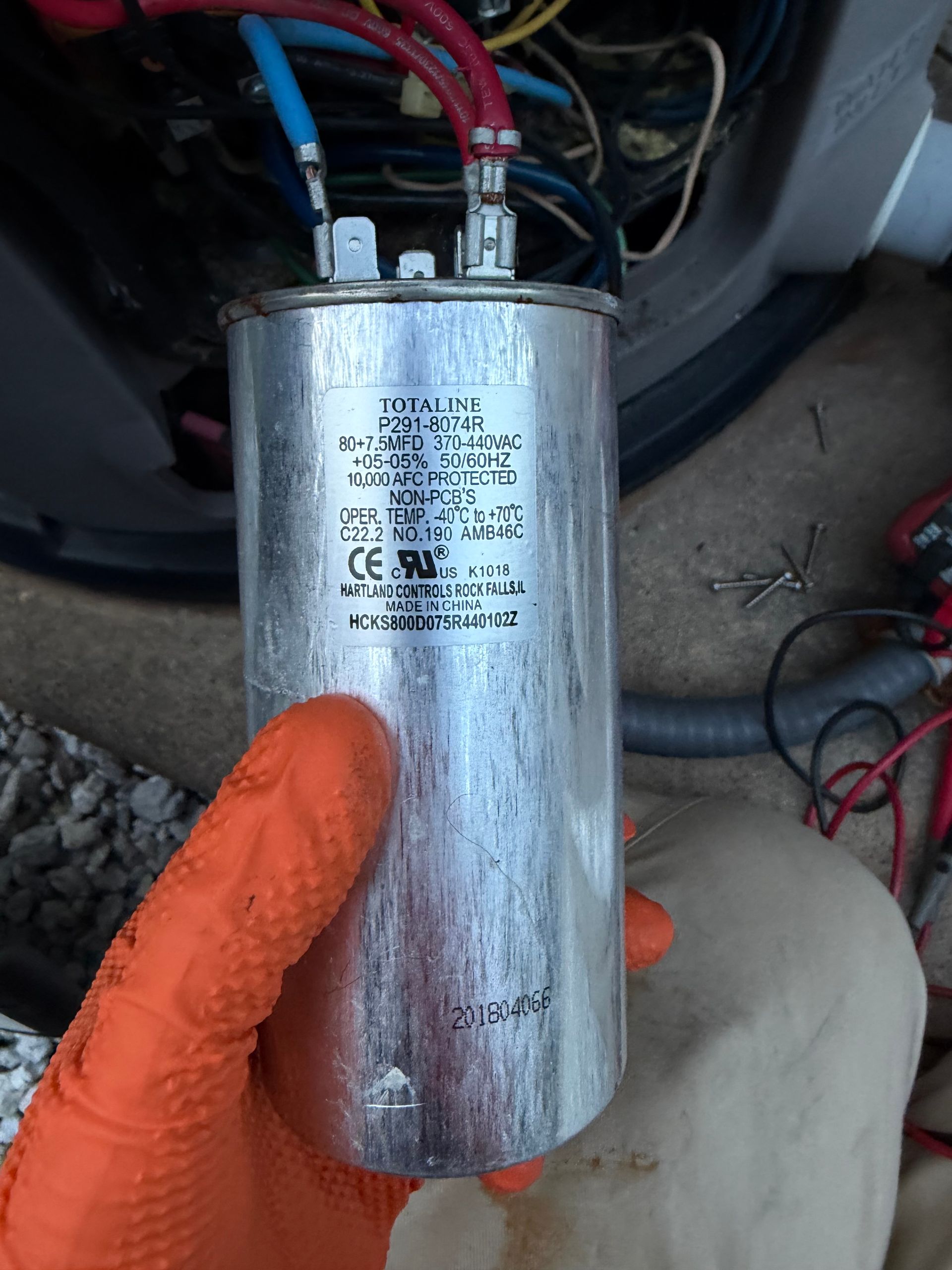 Hand holding a silver cylindrical capacitor with red and black wires attached in a vehicle engine bay
