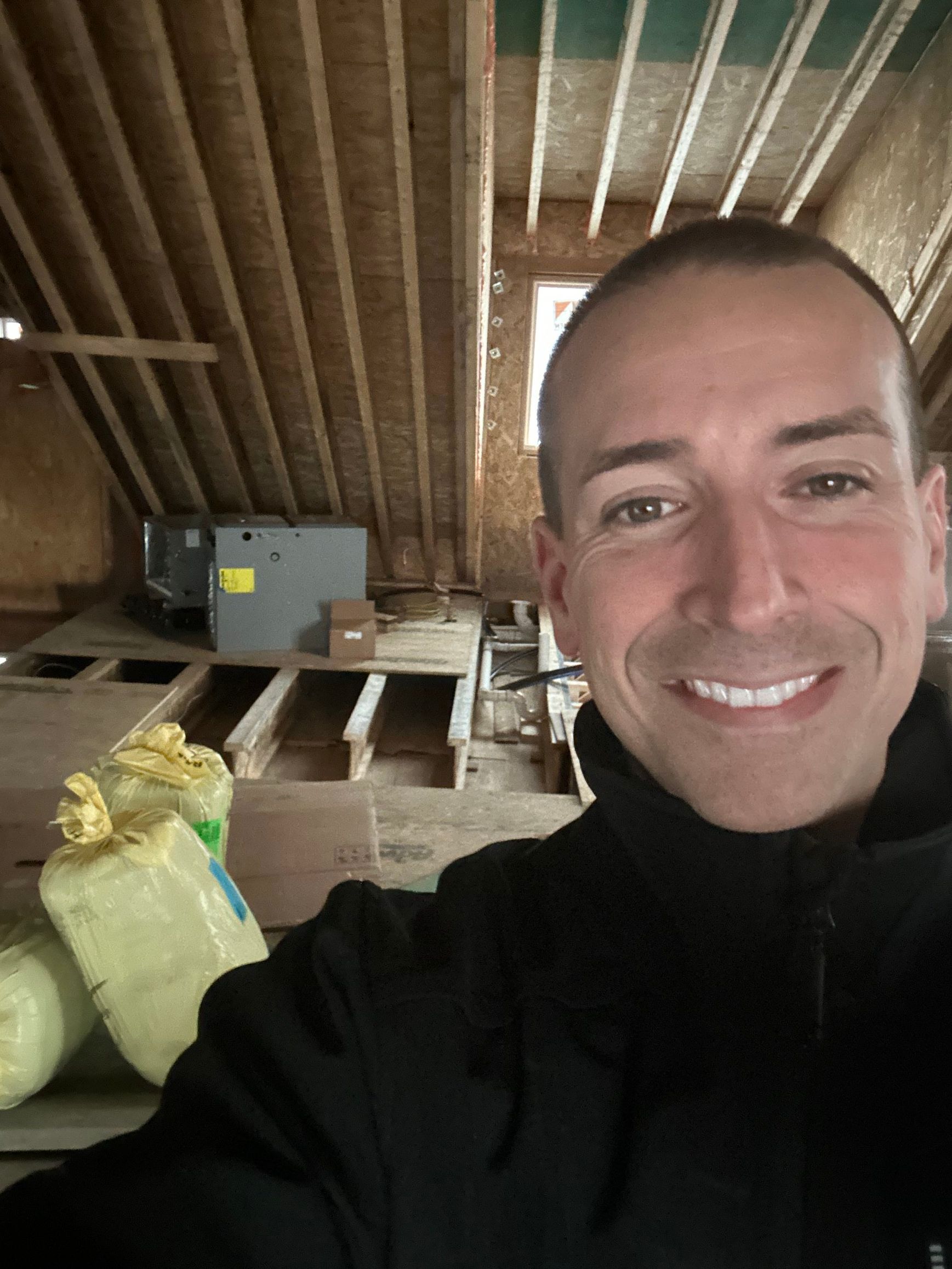 Man in black jacket smiles, taking a selfie in a construction setting; exposed beams, insulation, and HVAC unit visible.
