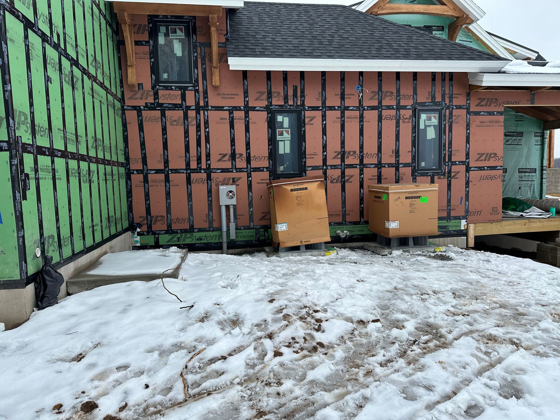 Construction site exterior. House with black windows and brown siding, covered in snow, boxes on the ground.