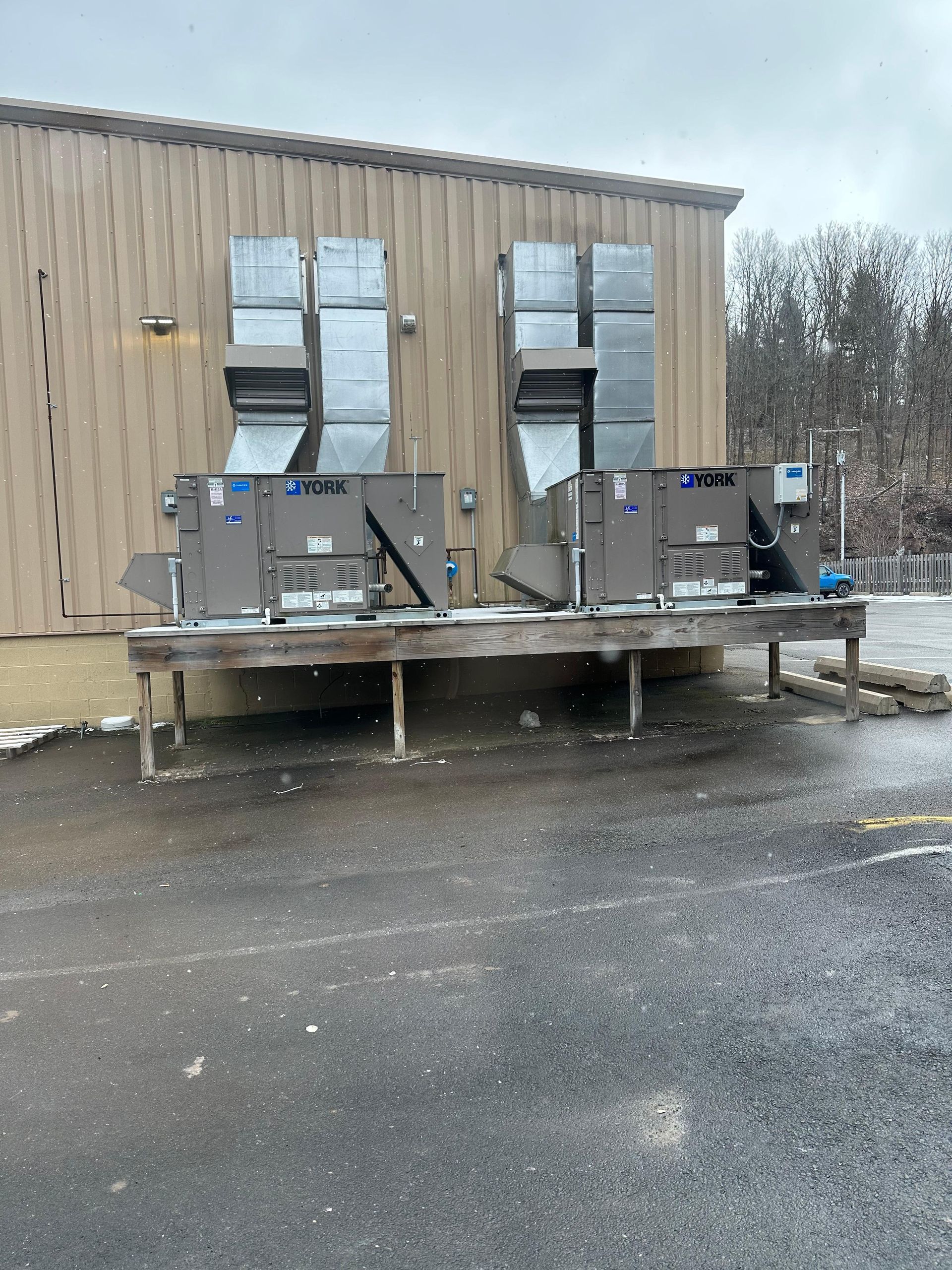 Two industrial HVAC units on a raised wooden platform against a tan building; snowing.