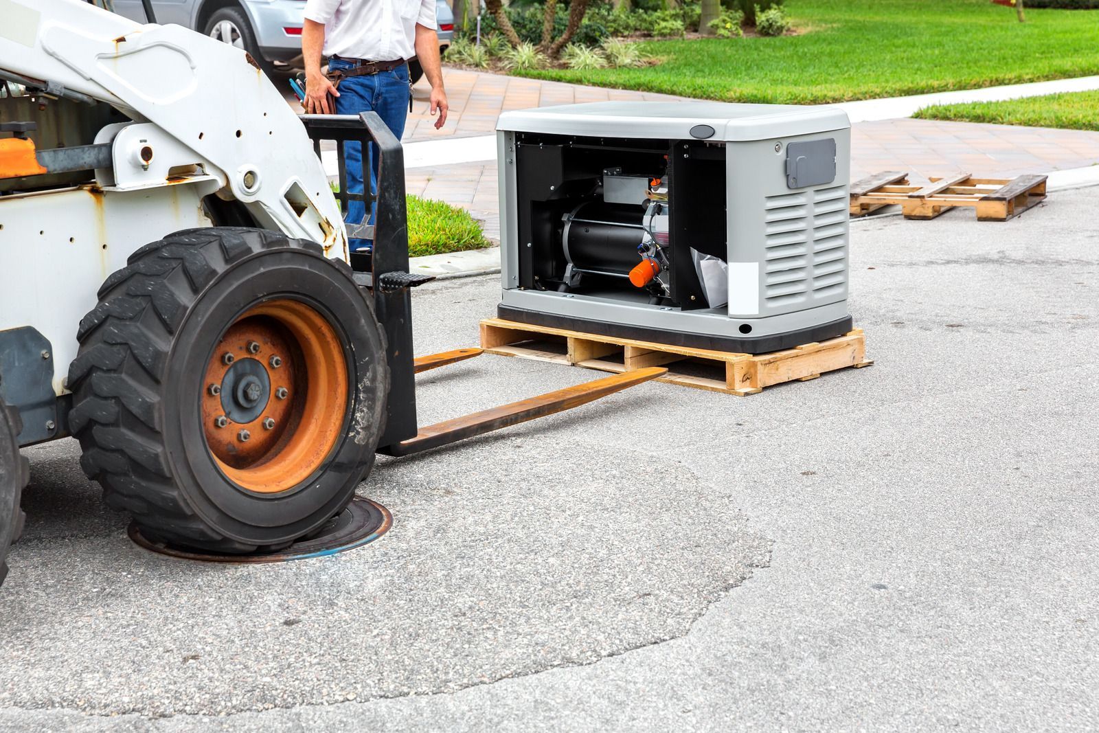 Skid steer forks lifting a generator on a pallet. A person stands nearby on a cracked asphalt street.