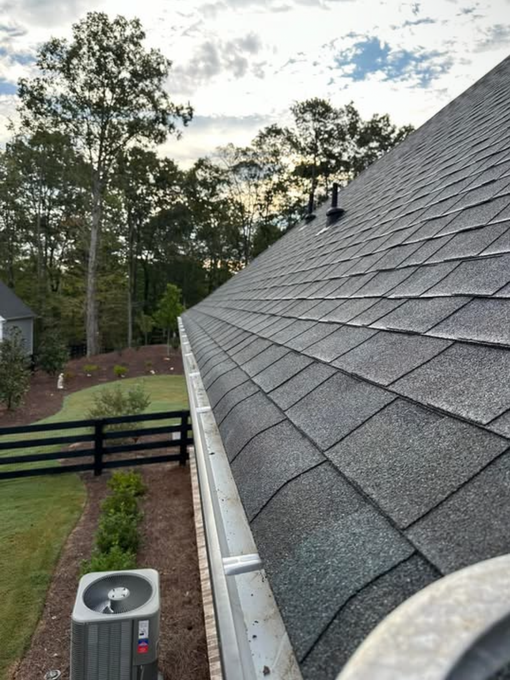 Pressure washed sloped gray shingle roof beside a yard with trees, fence, and an HVAC unit at dusk.