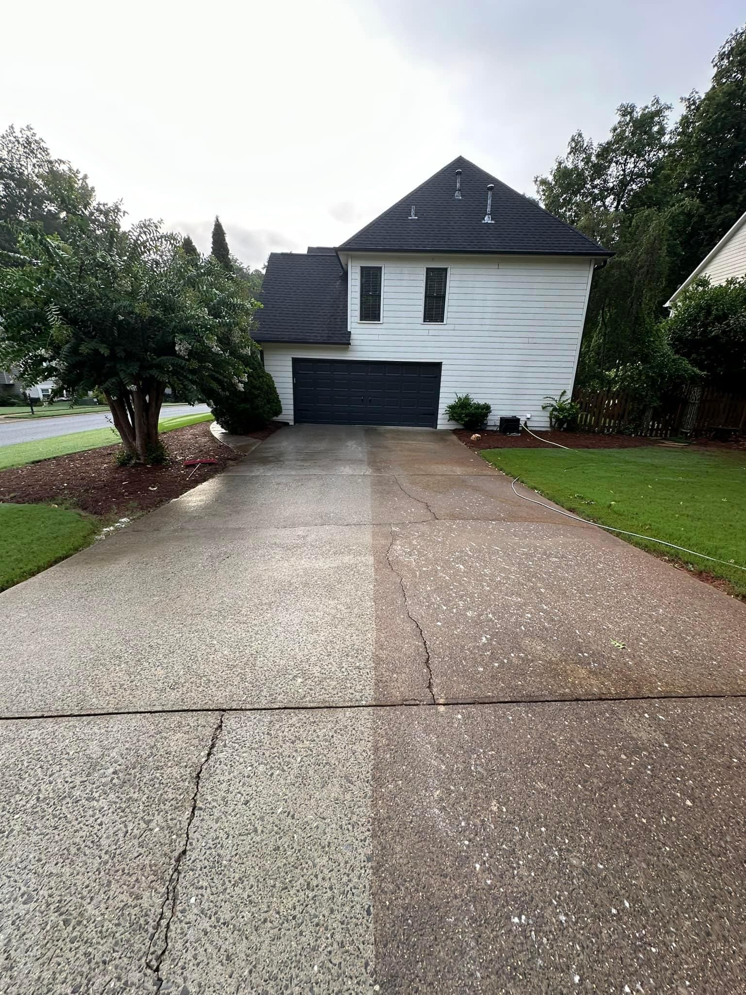 Pressure washed concrete driveway leading to a white two-story house with a dark roof and garage, framed by green lawns and trees.