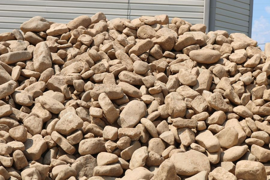A large pile of light brown, rounded stones against a grey siding wall.