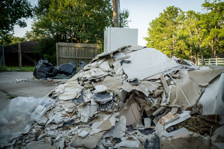 A large pile of construction debris, including drywall fragments and trash bags, sits outdoors near a wooden fence.