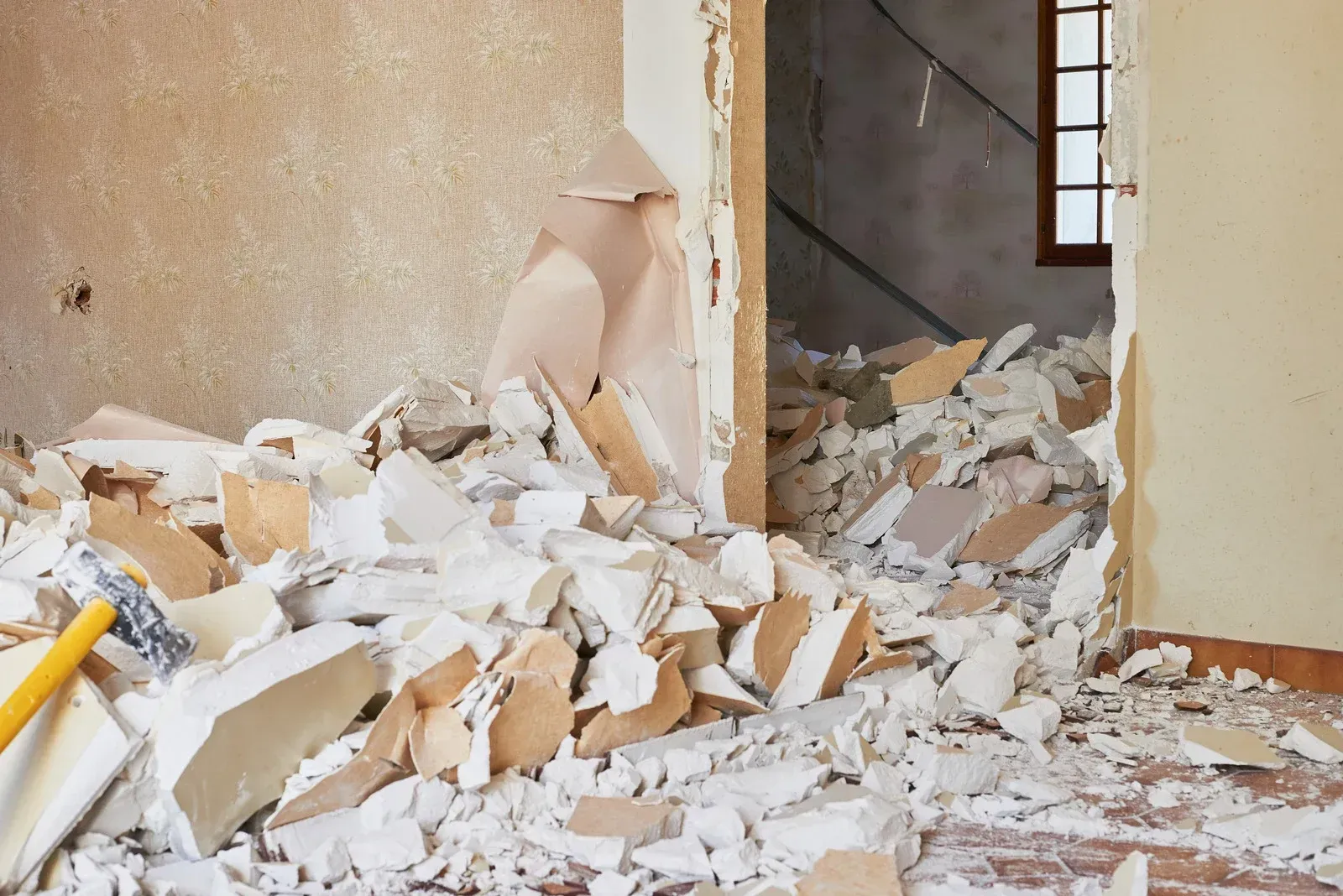 A room undergoing demolition with a large pile of broken drywall debris on the floor and a hammer in the foreground.