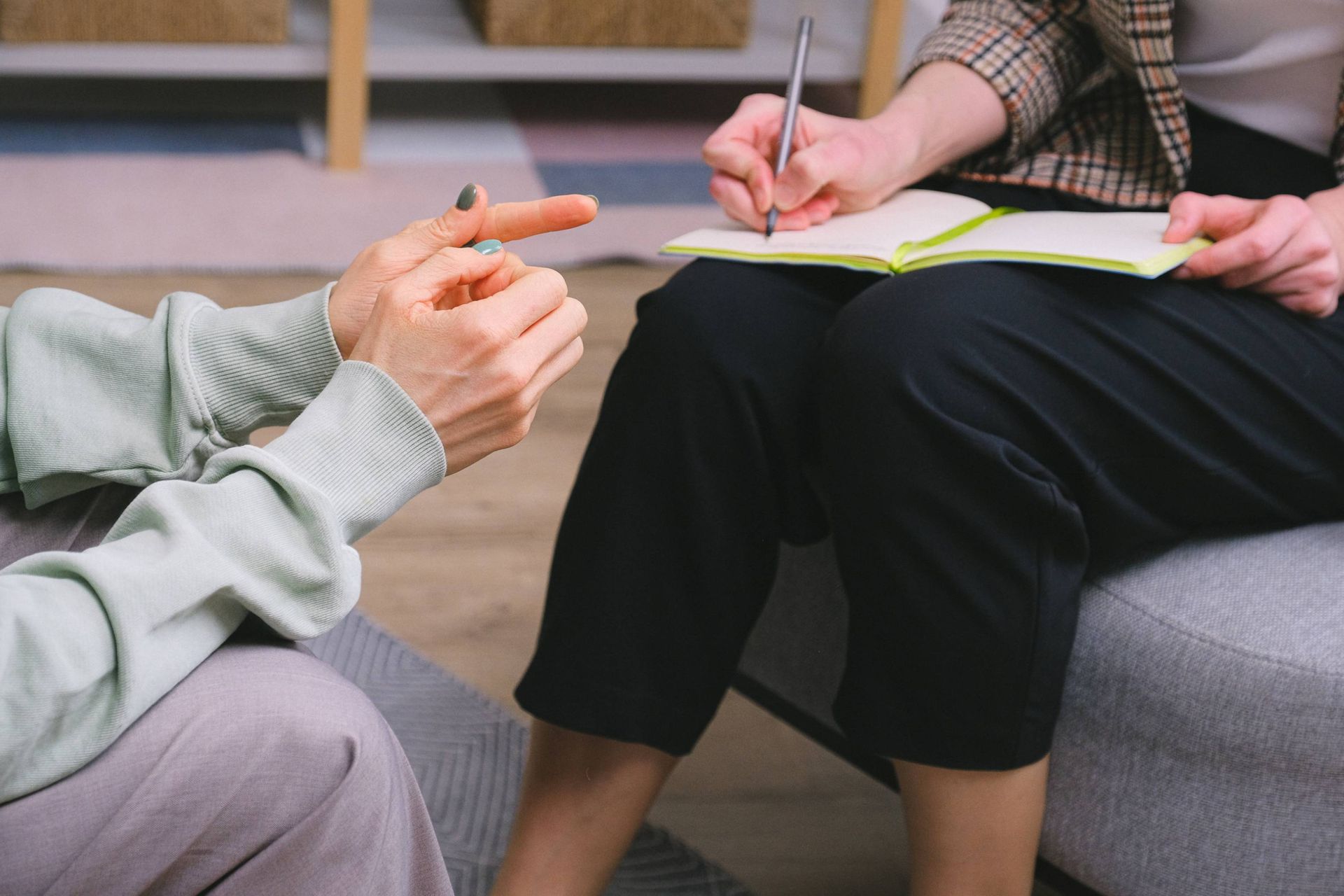 Person gestures while another takes notes in a notebook on a couch.