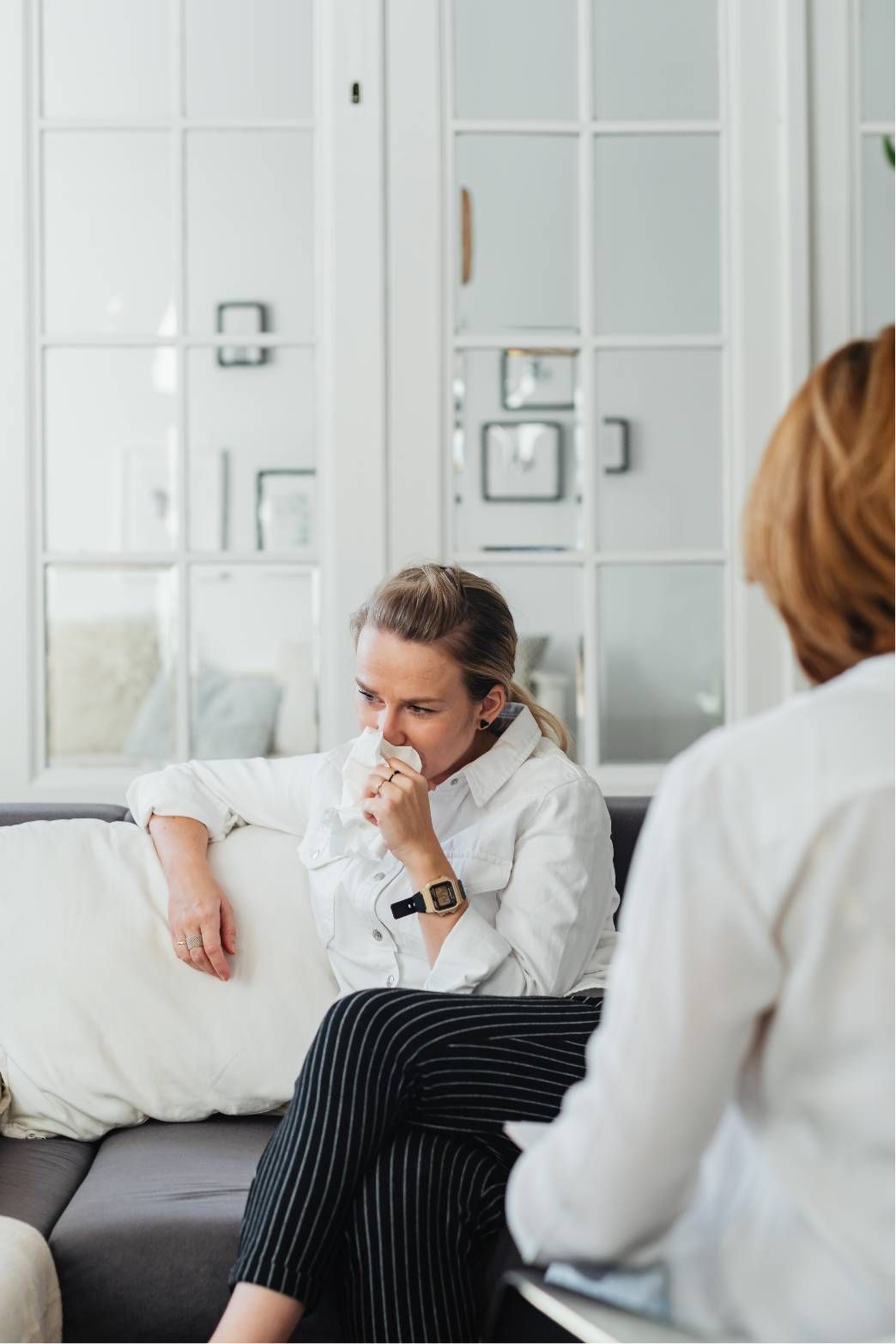 Woman in therapy, covering her mouth with tissue, seated on a couch; therapist facing her.