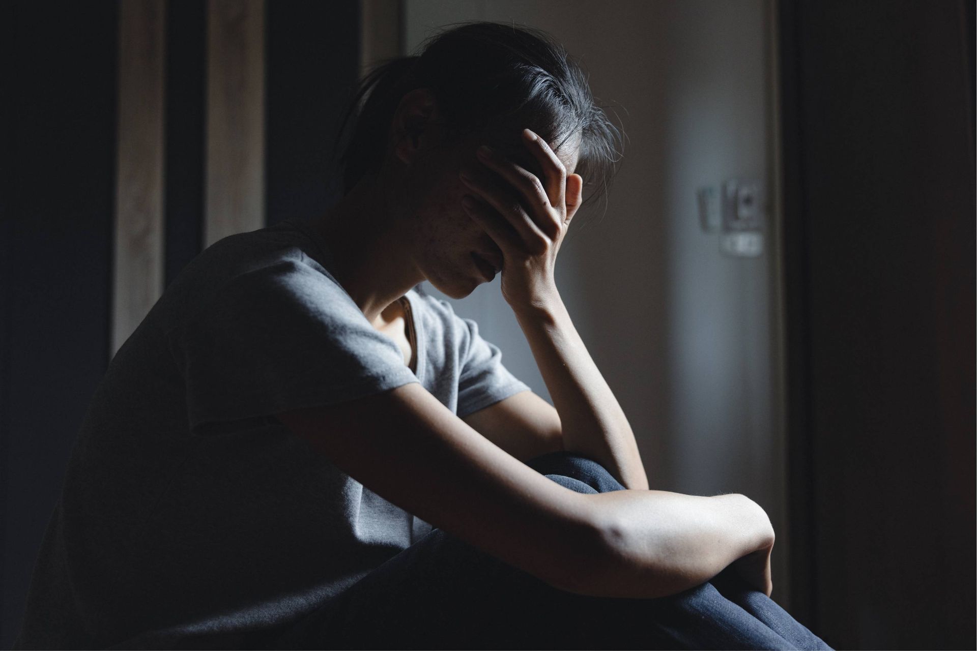 Person in a dimly lit room, seated with head in hands, appearing distressed.