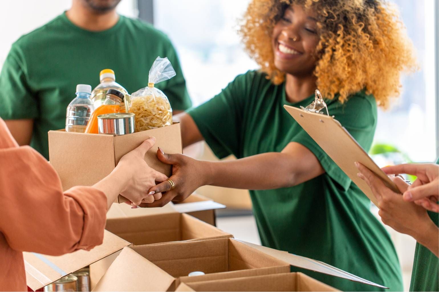 People in green shirts sorting food donations into boxes. Woman smiles while handing over a box.