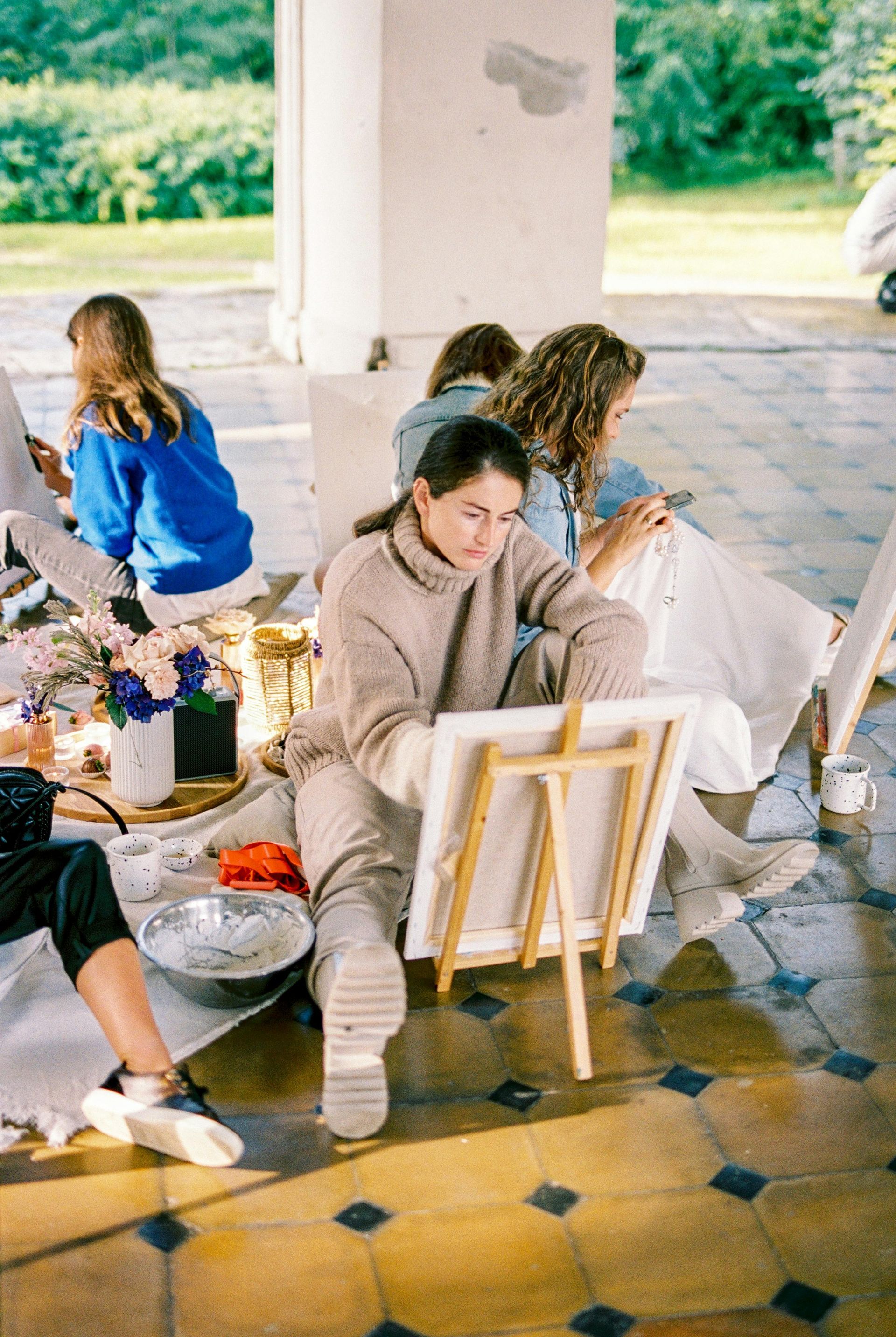 People painting outdoors at easels, sitting on a tiled floor. One person in beige sweater draws on canvas.