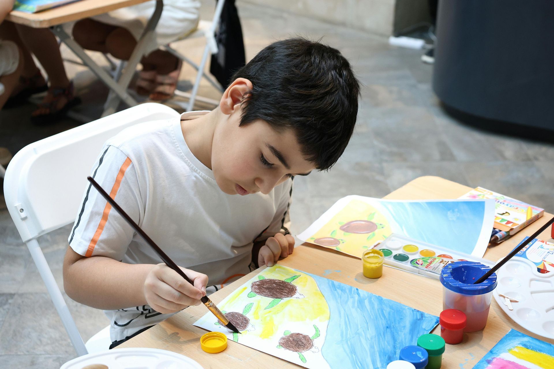 Boy focused on painting with watercolors at a table. Paints and artwork are visible.