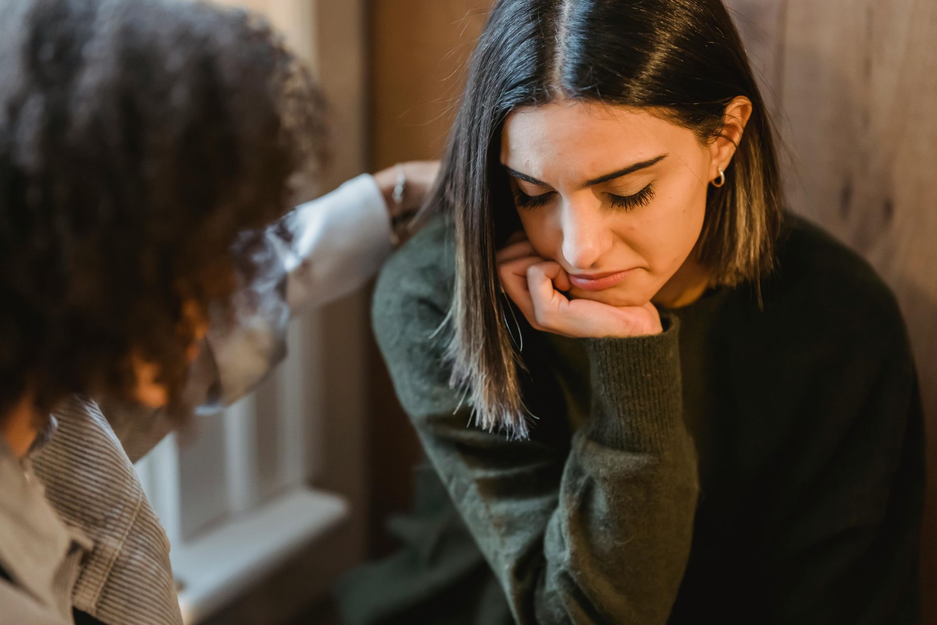 Woman with head in hand looks down, comforted by a person's hand on her shoulder. 