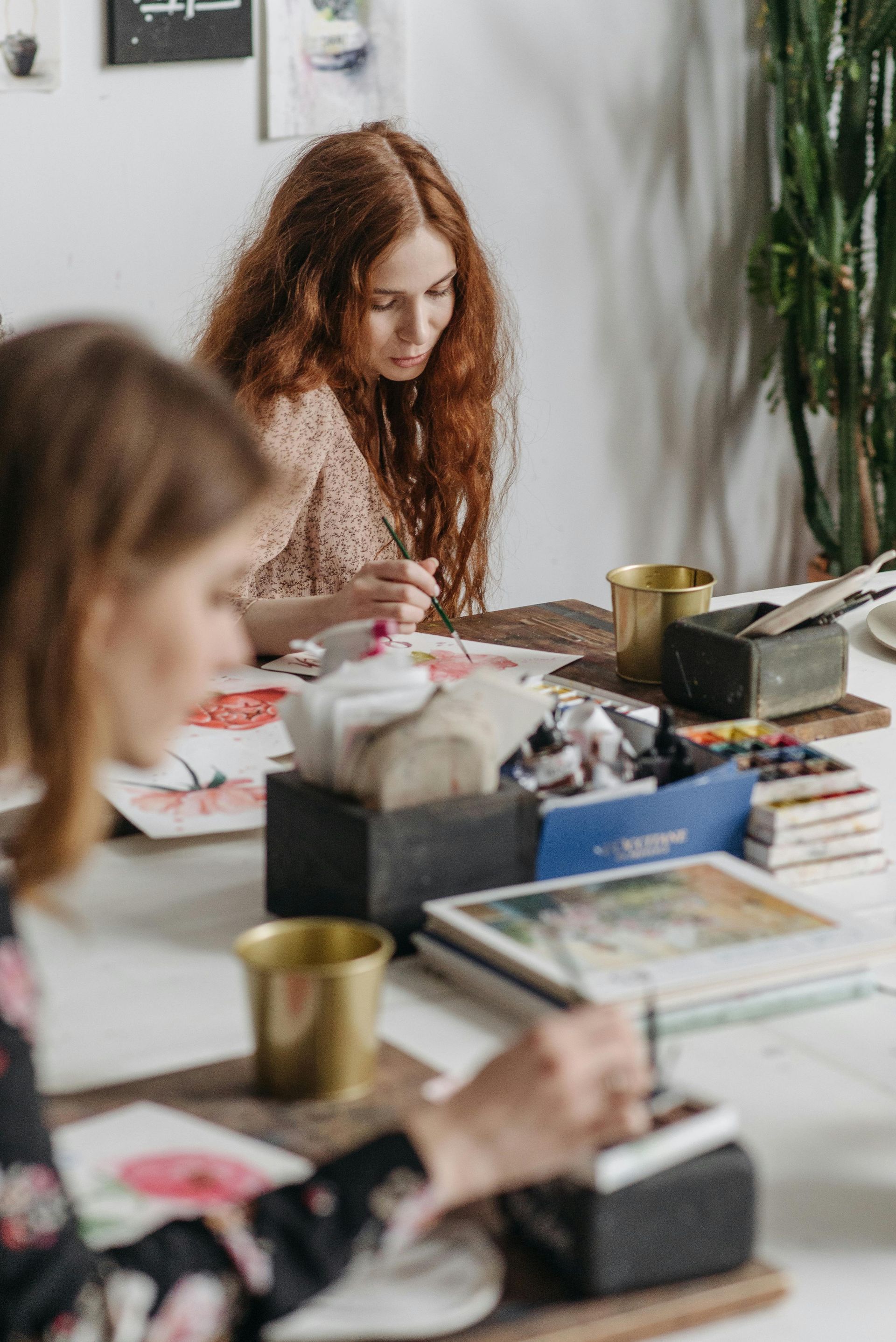 Two people painting at a table; one is concentrates on her work, paints, art supplies present.