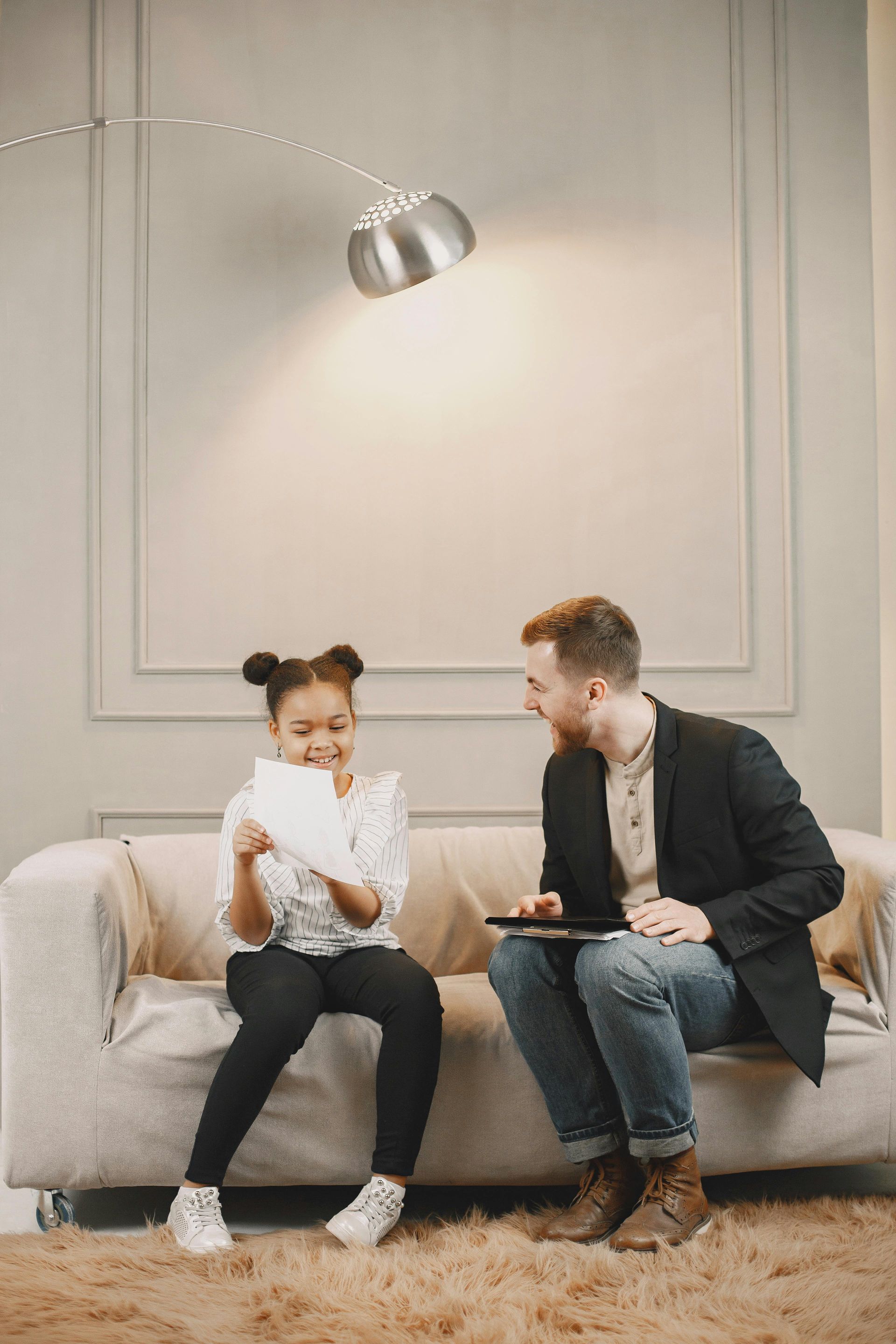 Girl on couch looking at paper; man smiles, holds tablet; near floor lamp.
