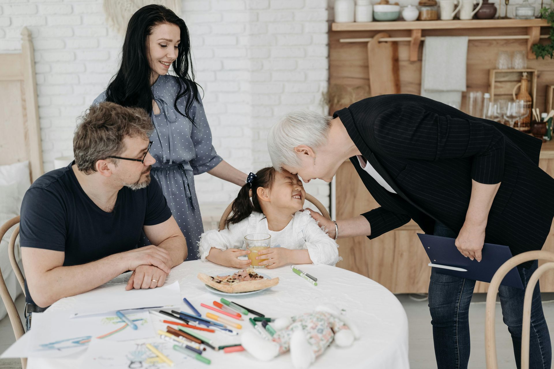 Family gathered around a table. A woman kisses a child's forehead. Others smile. Colorful drawing supplies on the table.
