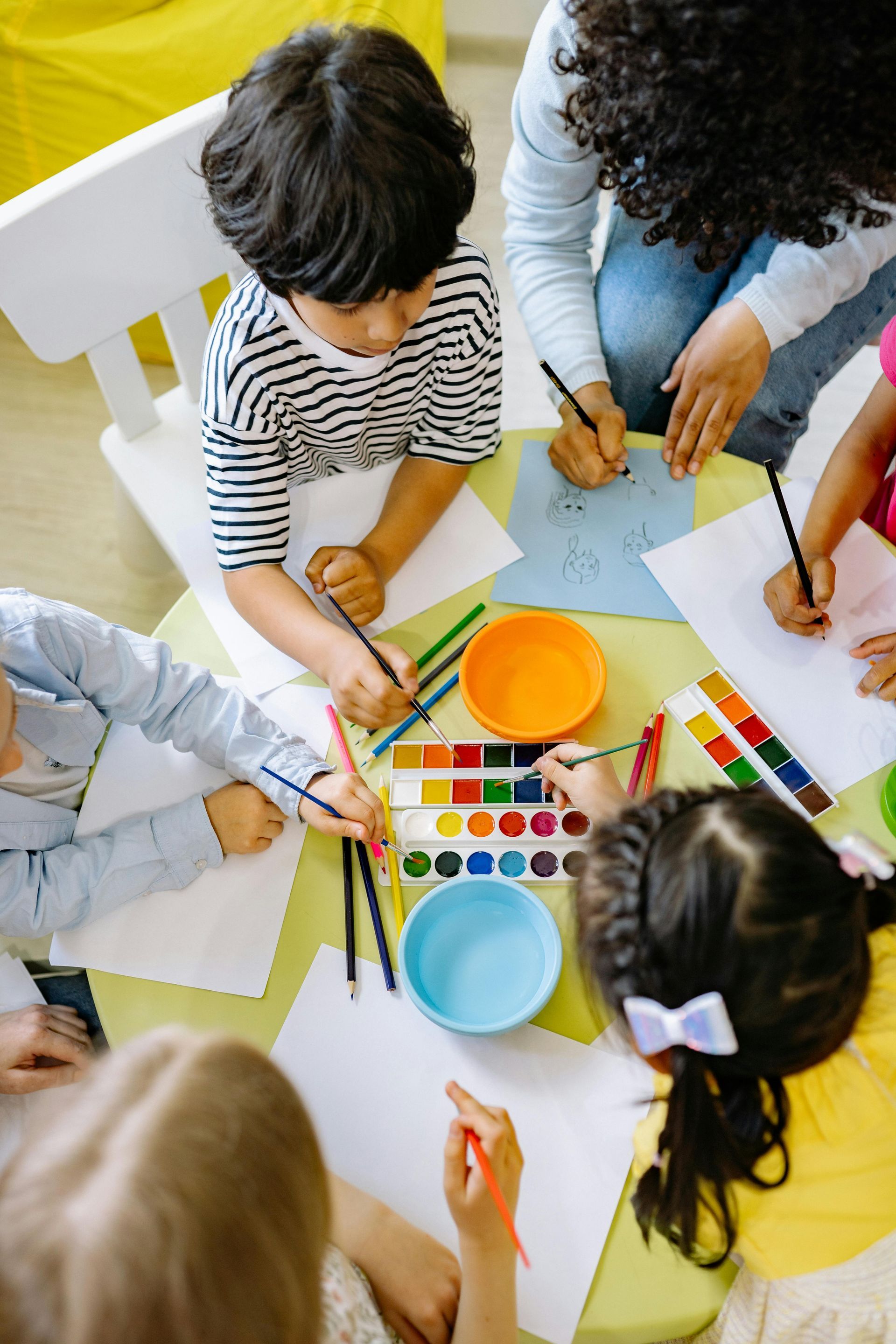 Children and an adult draw at a table with paints and paper.