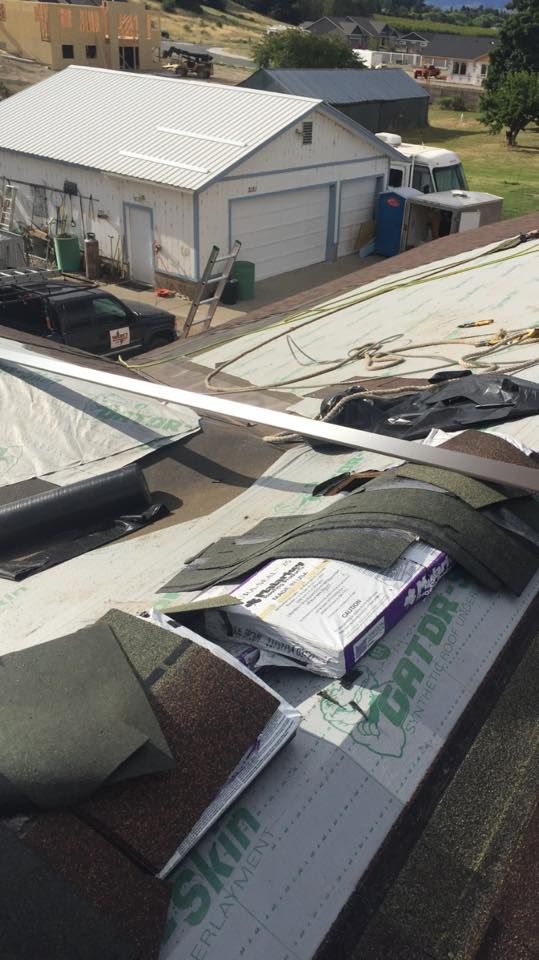 Roofing materials scattered on a damaged house roof beside a white shed in a residential yard.