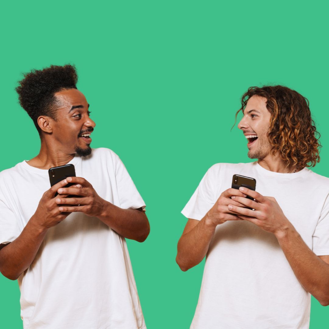 Two people in white shirts smiling at each other while holding smartphones against a green background