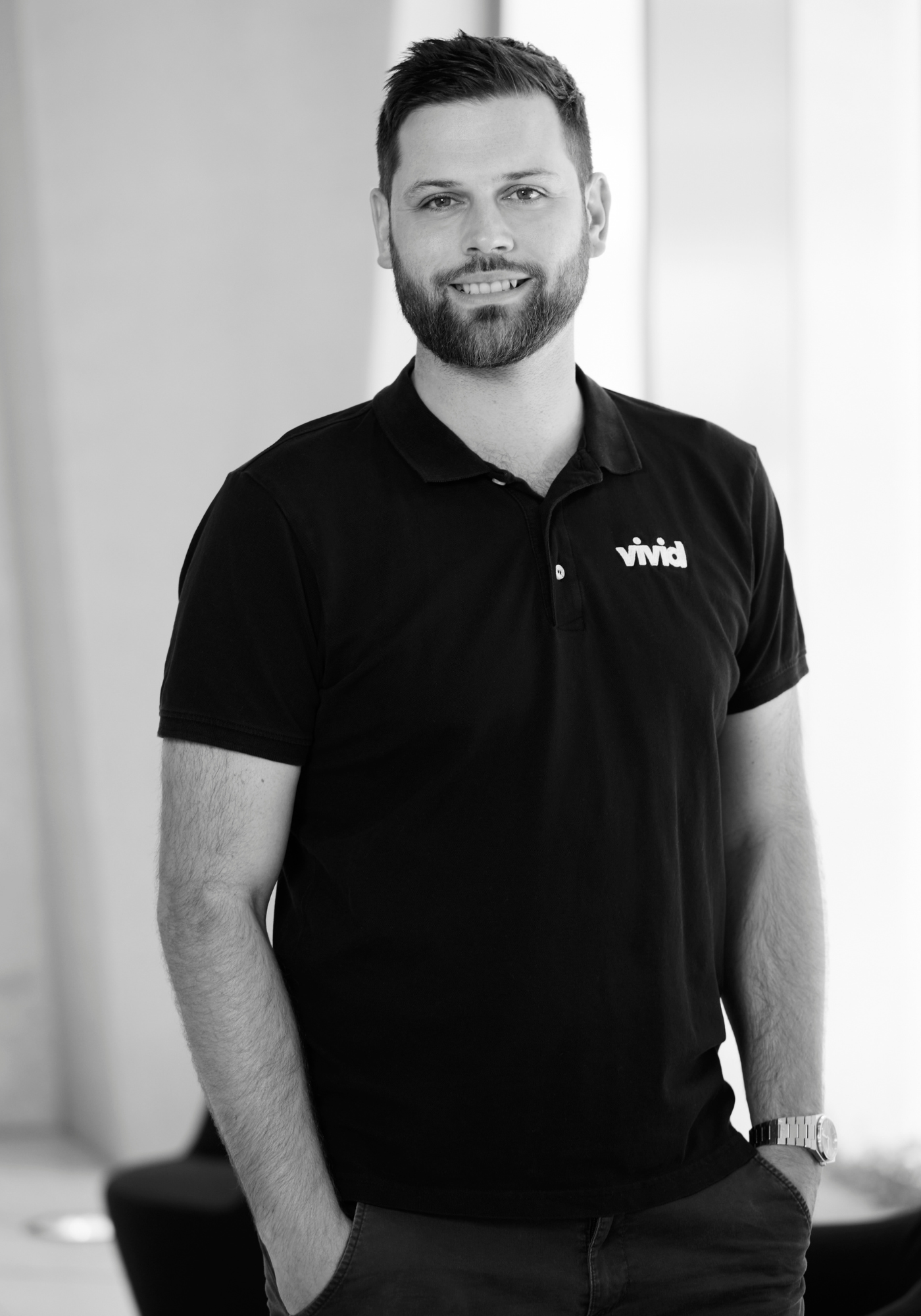 Man standing in a black polo shirt, smiling in a black-and-white portrait.