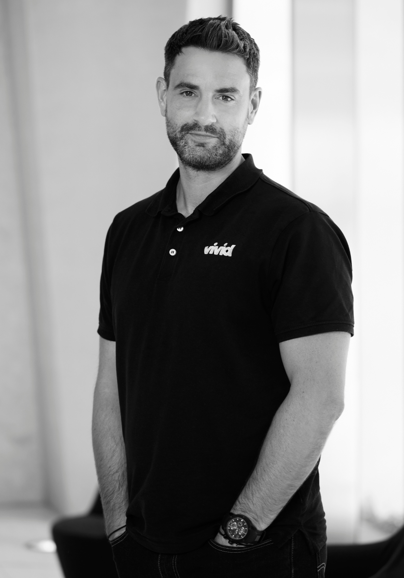Man standing indoors in a black polo shirt and watch, facing the camera with hands in pockets.