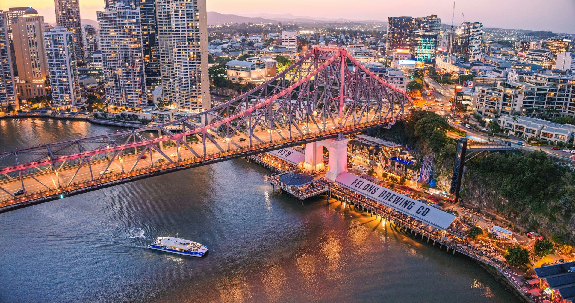 City bridge lit pink over a river at dusk, with boats and high-rise buildings in the background.