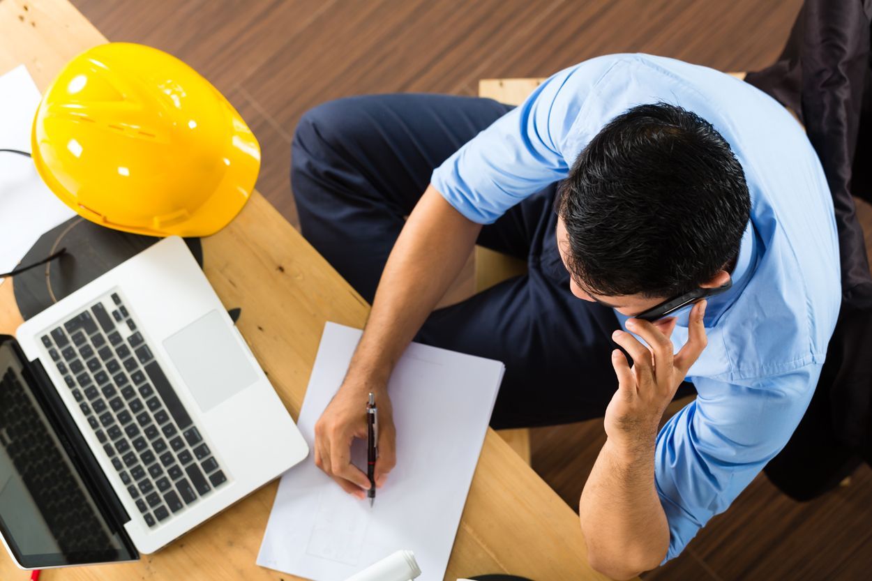 Man seated at a desk, wearing a blue shirt, talking on the phone, taking notes, with a laptop and hard hat nearby.